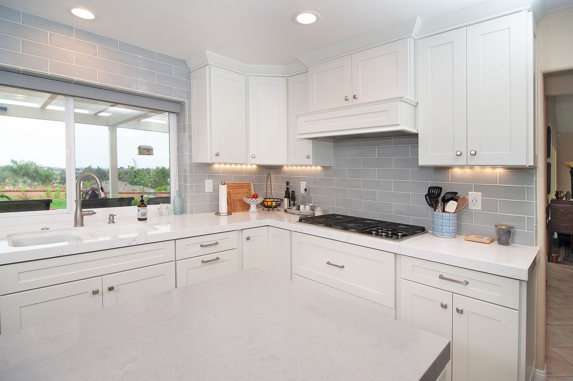 White kitchen with light gray backsplash and countertops, and a large window looking outdoors.