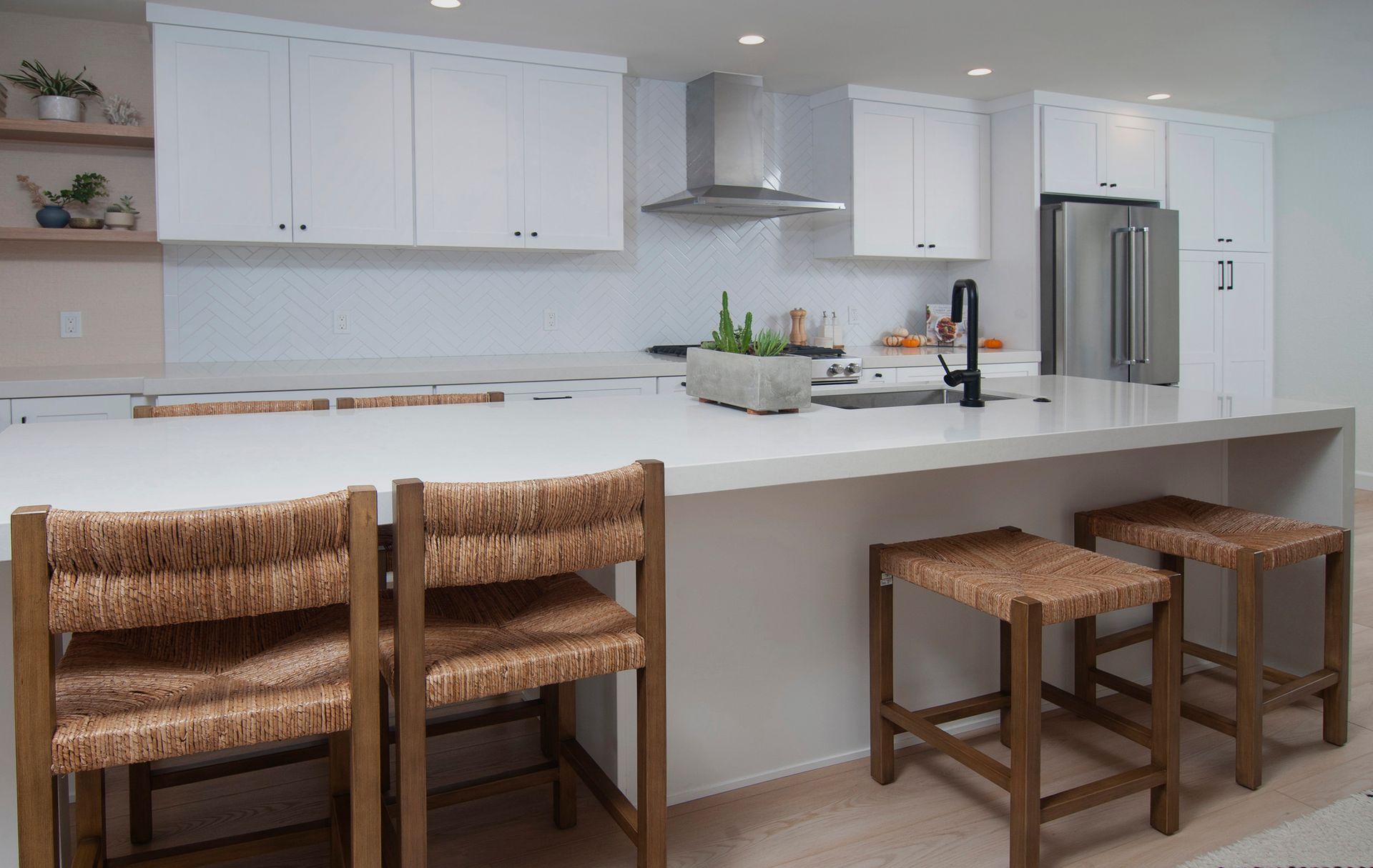 Modern white kitchen with island, three woven stools, stainless steel appliances, and white countertops.