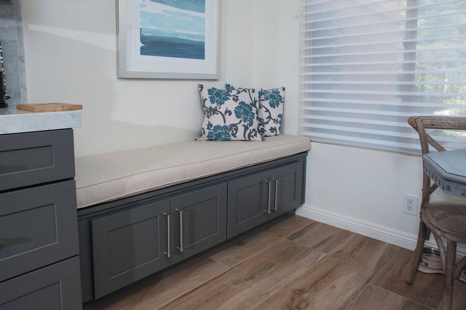 A built-in gray bench with cream cushion and floral pillow by a window with wood flooring.