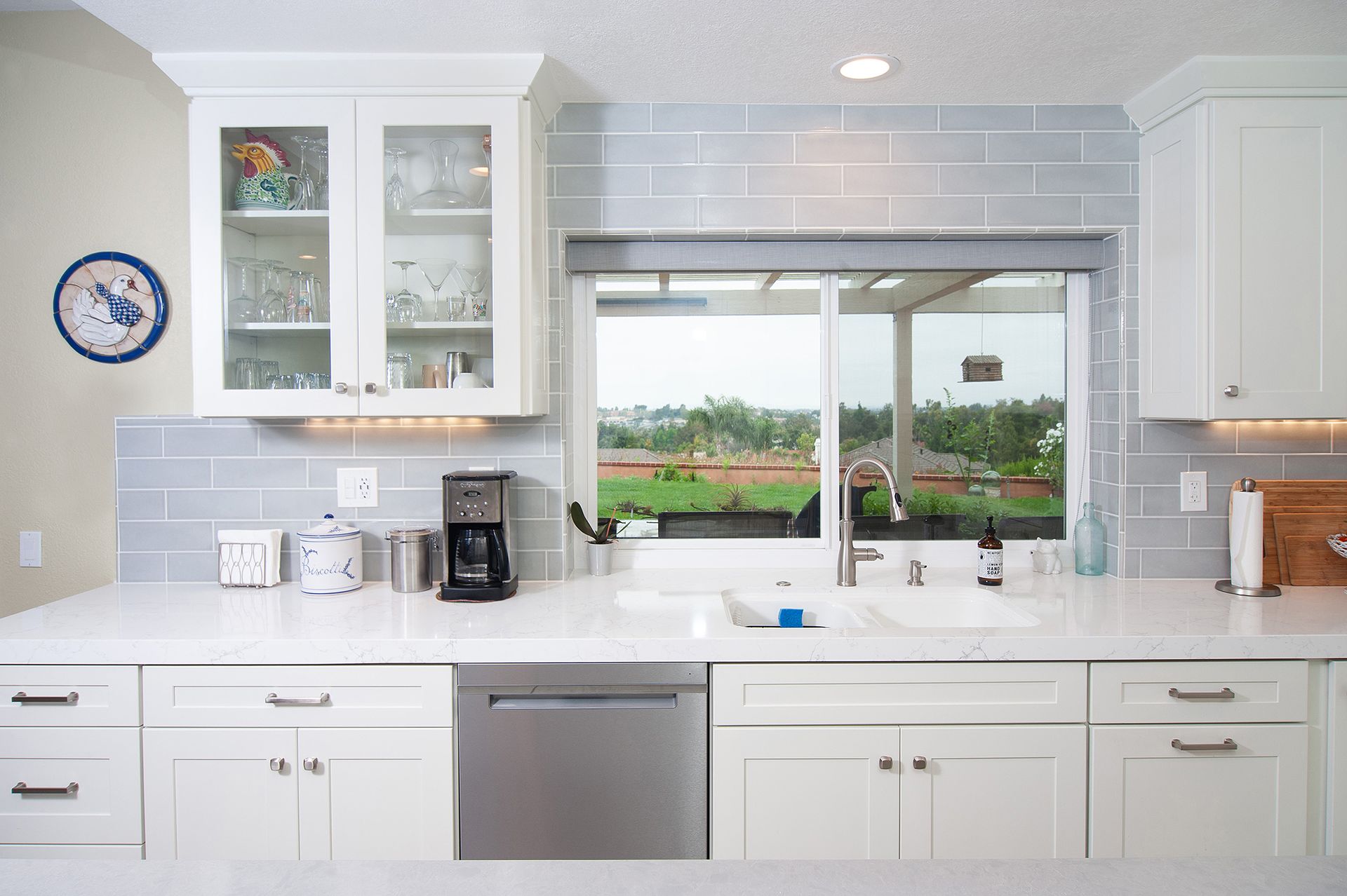 White kitchen with a light blue tile backsplash and a window overlooking a green outdoor space.