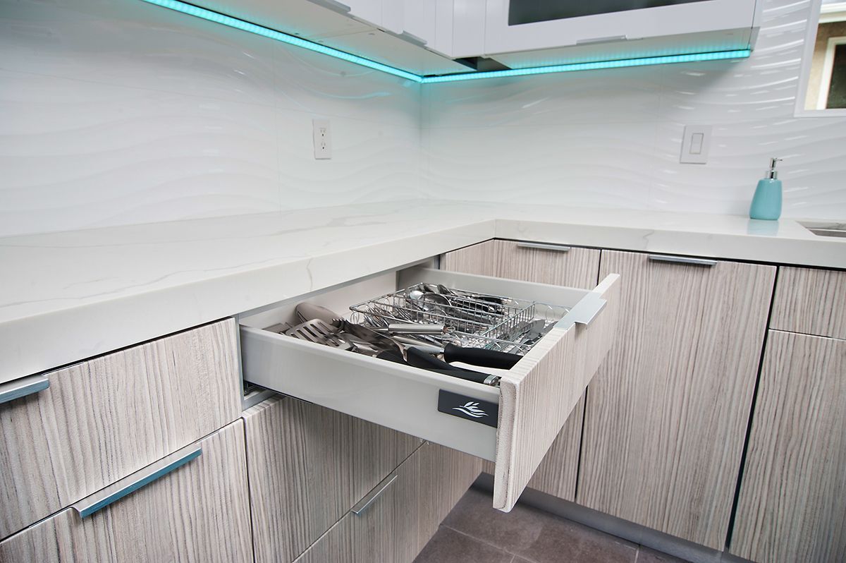 Open kitchen drawer with silverware, light-colored wood cabinets, and under-cabinet lighting.