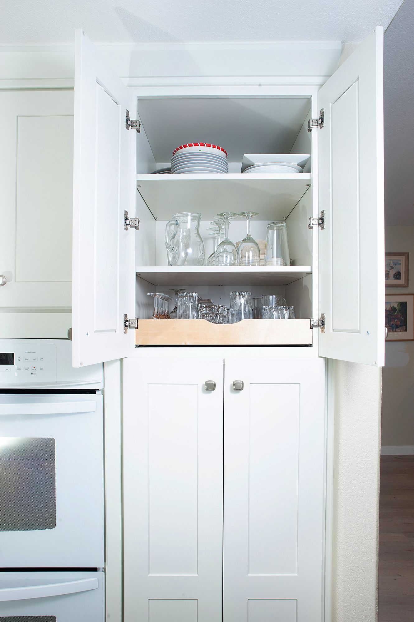 White kitchen cabinet open, revealing glassware on shelves, below an upper shelf with a dish.