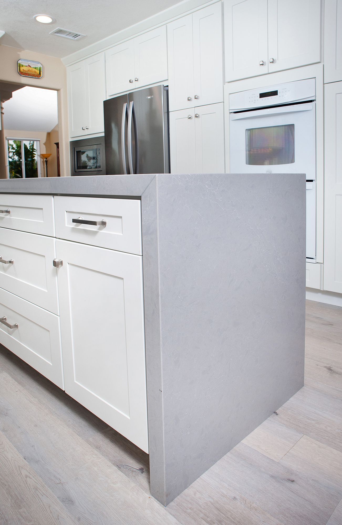 White kitchen island with gray countertop, white cabinets, stainless steel appliances, and light wood floor.