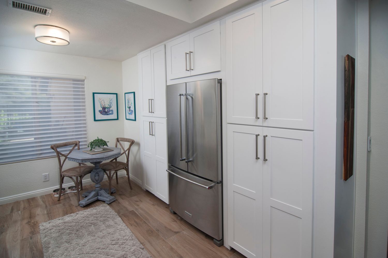 White kitchen with stainless steel refrigerator, tall cabinets, small dining table, and wood-look flooring.