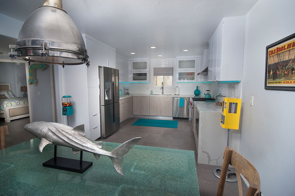 Modern kitchen with a large silver shark sculpture on a turquoise table, stainless steel appliances, and light blue accents.