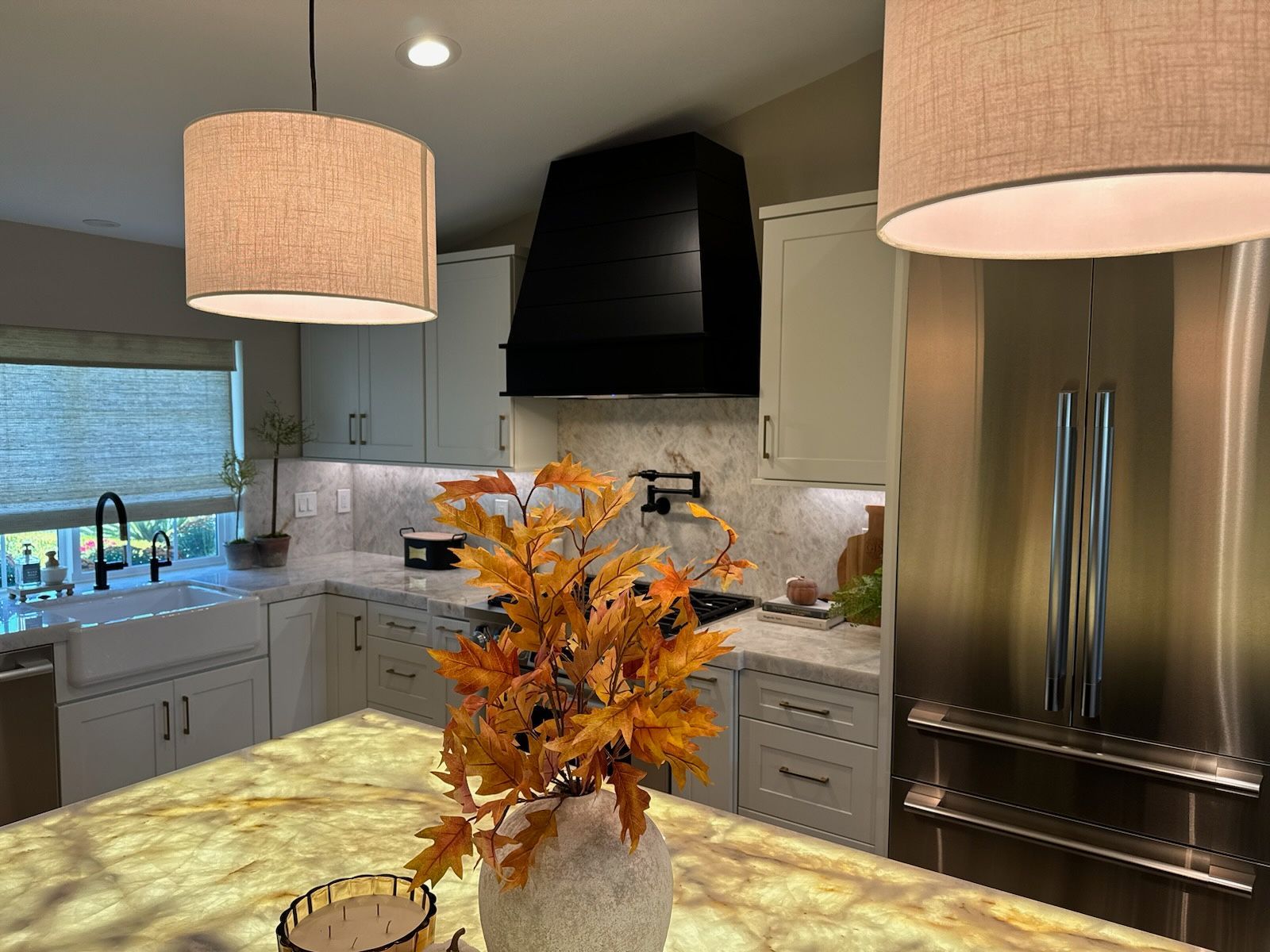 Cozy kitchen with neutral cabinets, black range hood, island with autumn flowers, and linen drum pendant lights.