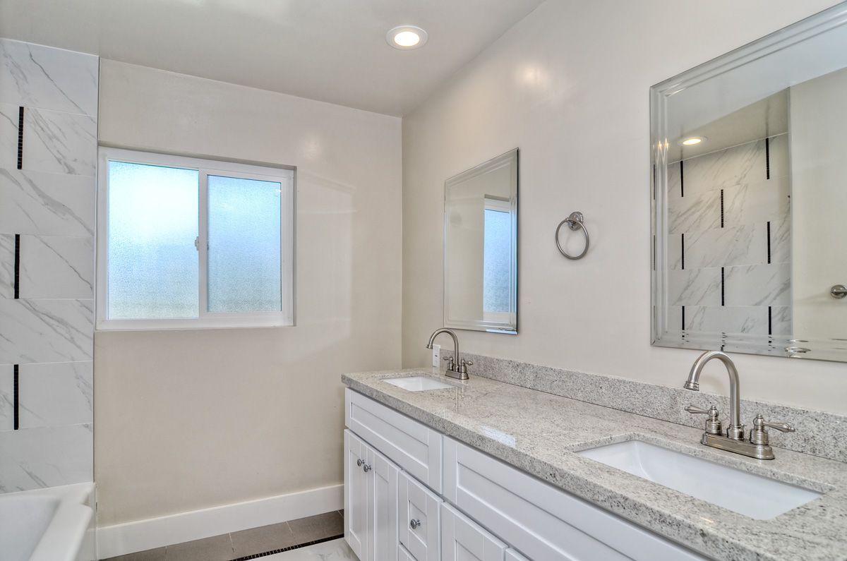 Bathroom with dual sinks, white cabinets, granite countertop, mirrors, and a frosted window.