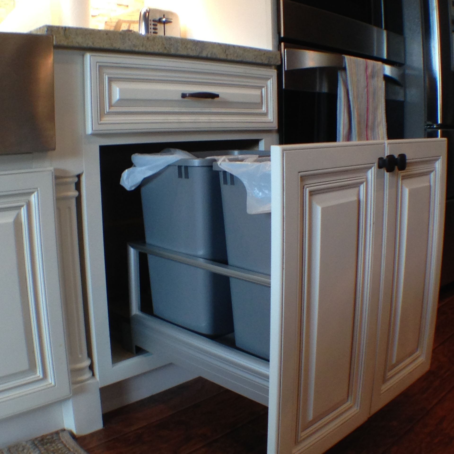White kitchen cabinet with pull-out trash bins. Gray bins, granite countertop, black hardware.