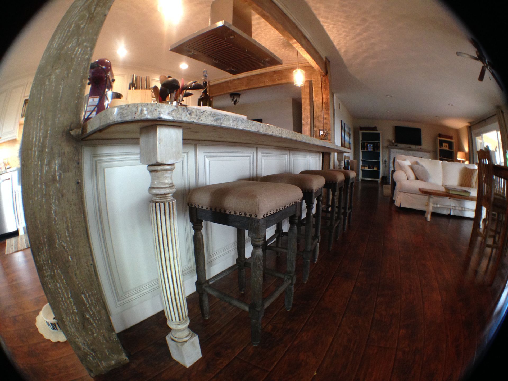 Kitchen with bar stools and living area in the background; wooden floors and light-colored cabinets.