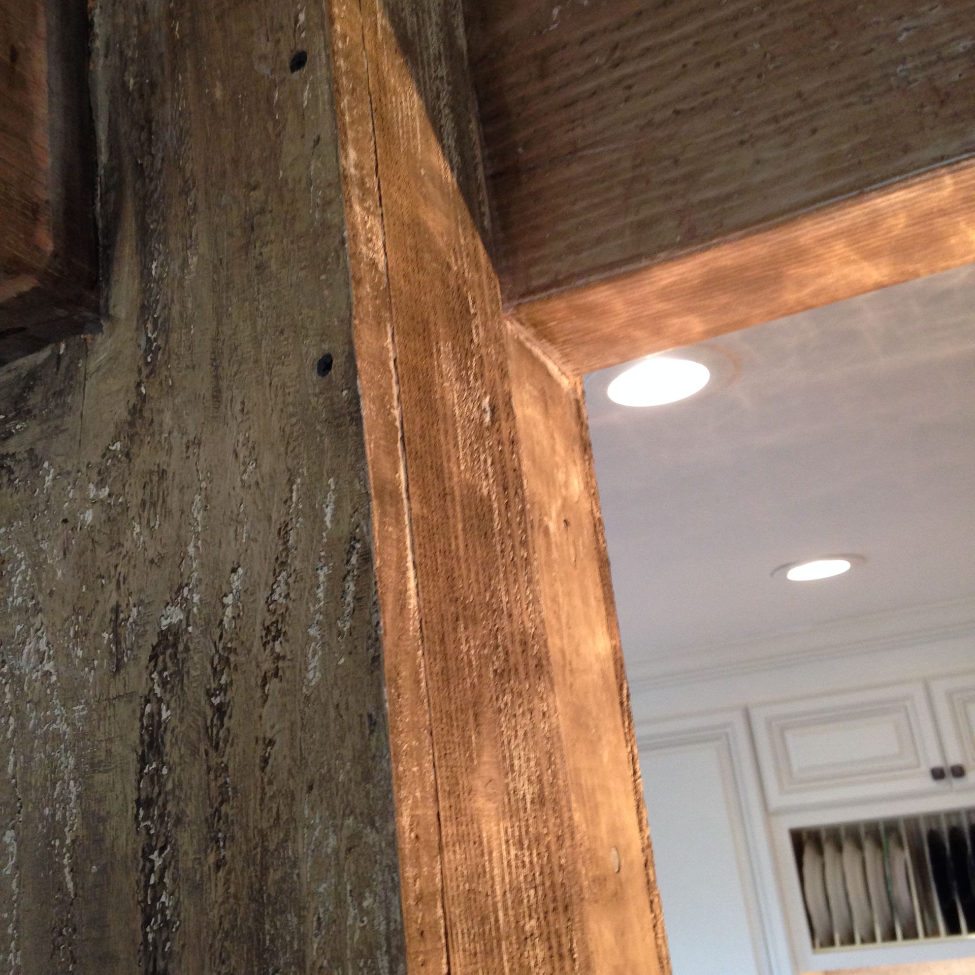 Wooden beams, ceiling and doorway frame a kitchen with recessed lights and white cabinets.