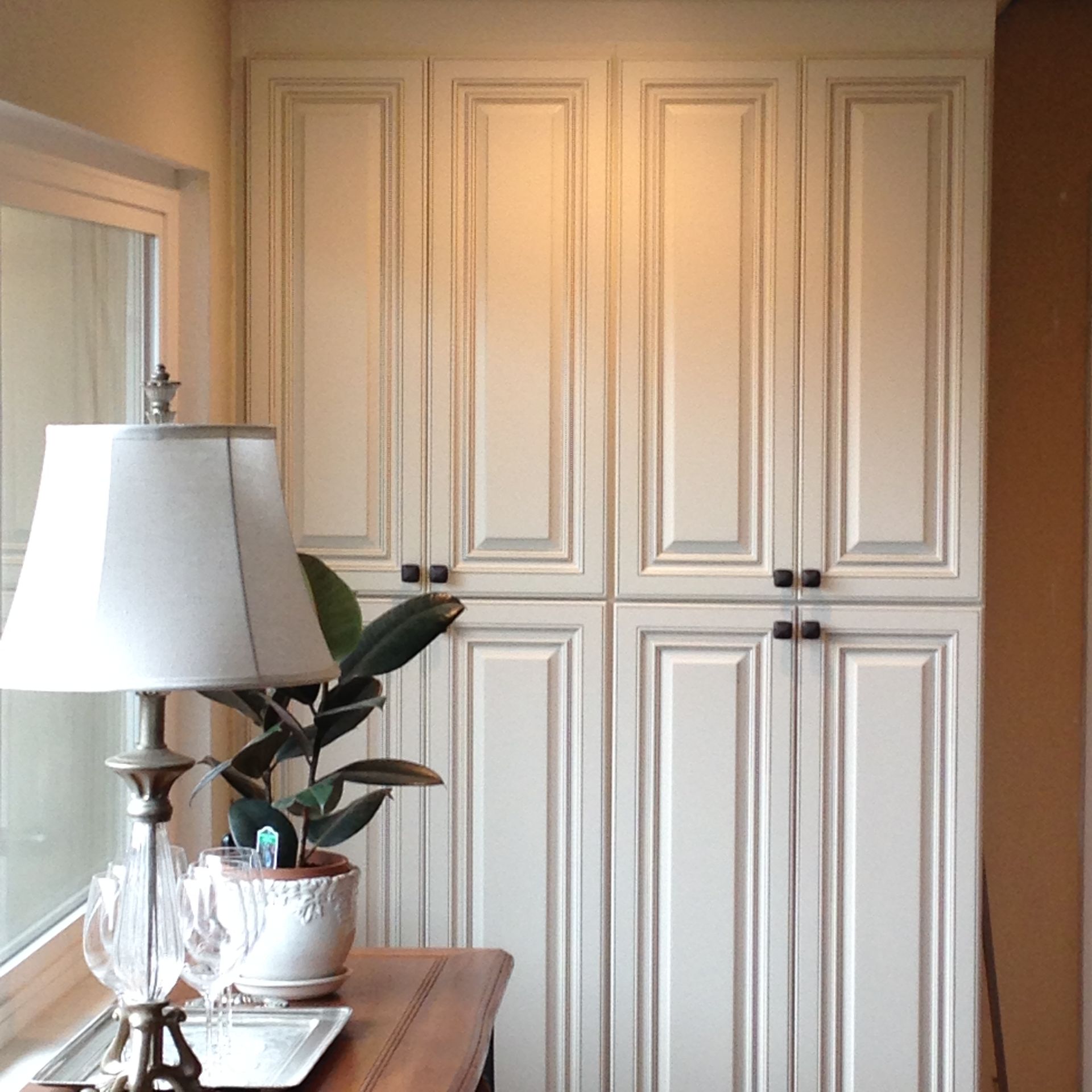 Cream-colored cabinets with black handles sit against a wall. A lamp and potted plant are on a table nearby.