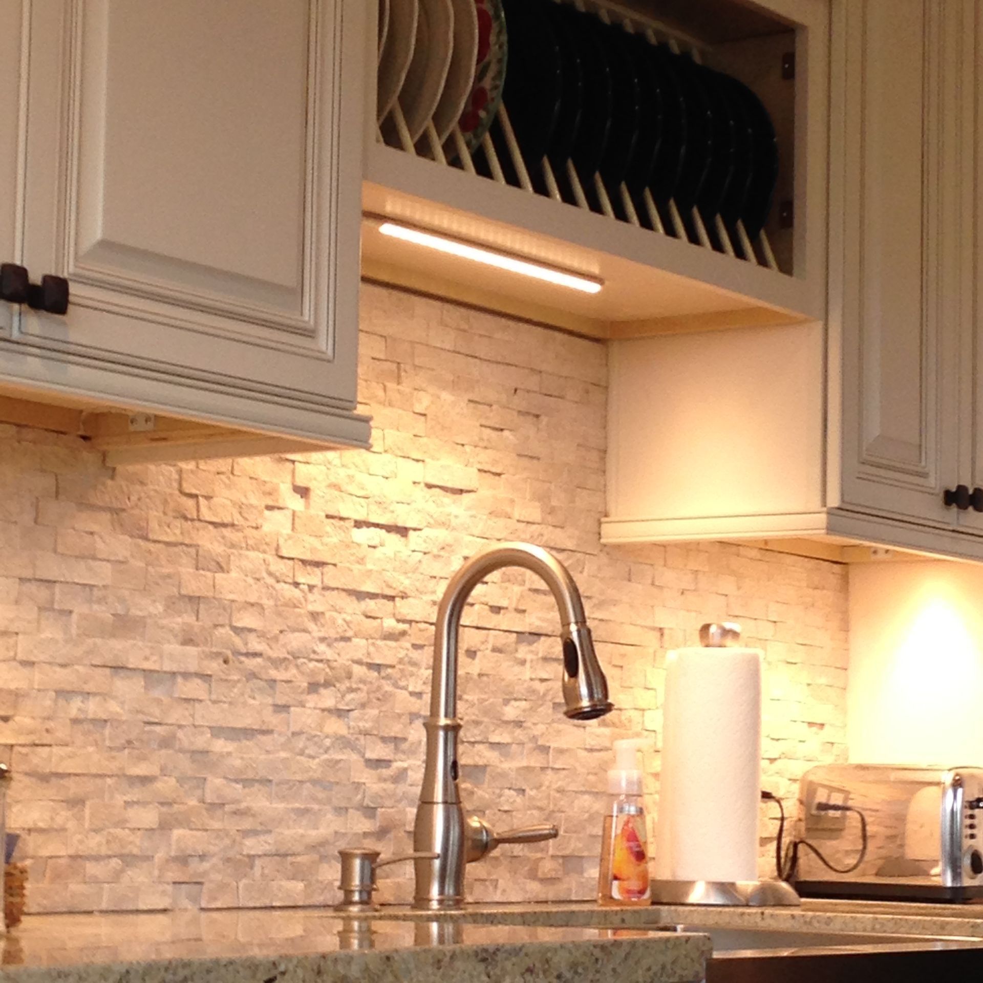 Kitchen with sink, faucet, and backsplash lit by under-cabinet lighting; dish rack above sink.