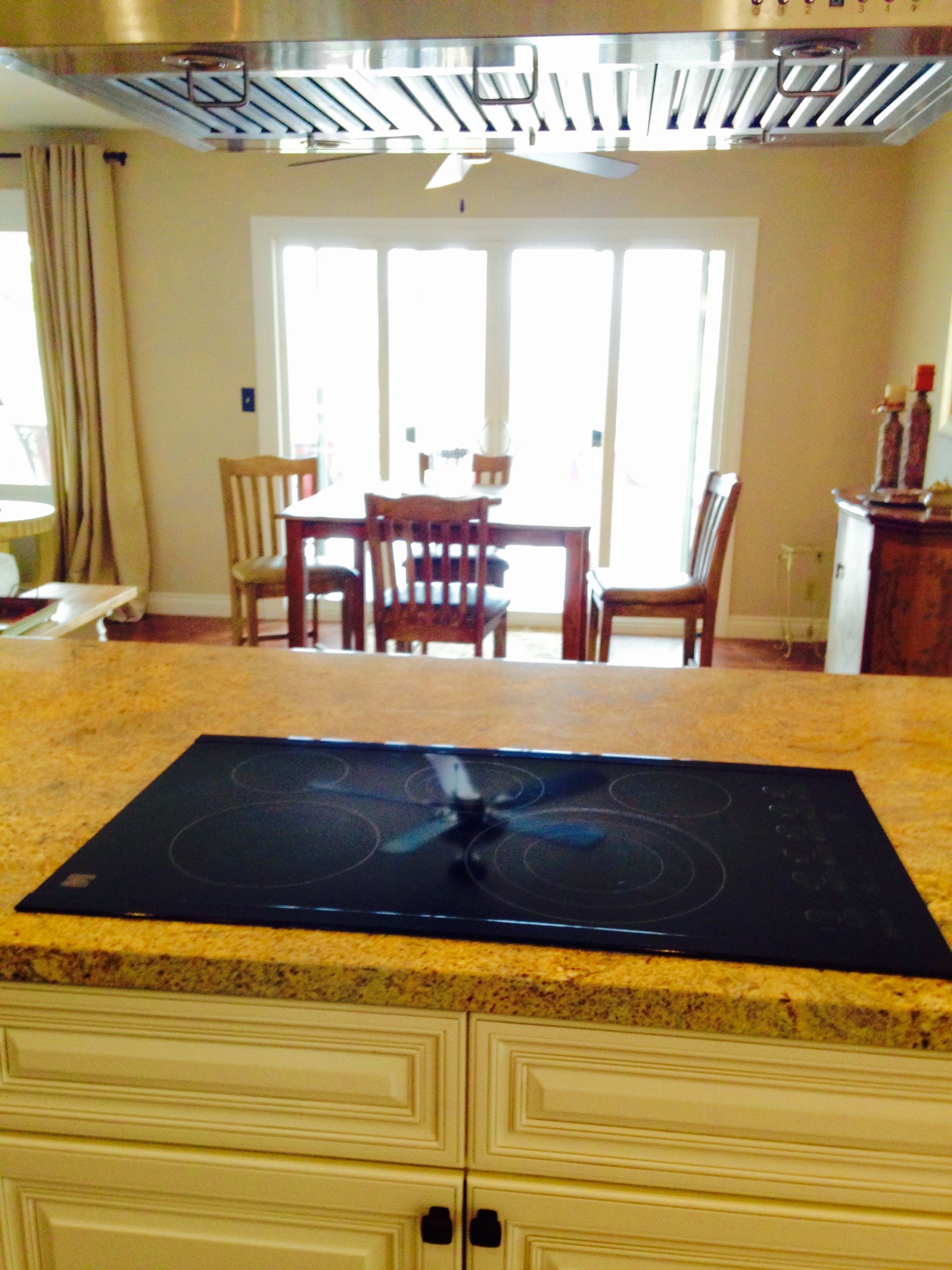 Kitchen with a stovetop on a granite island, looking towards a dining table by a window.