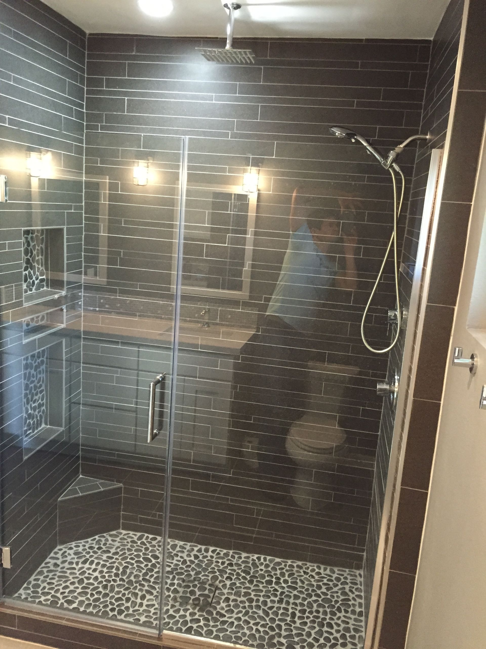 Modern dark-tiled shower with a glass door, pebble floor, and a vanity in the background.