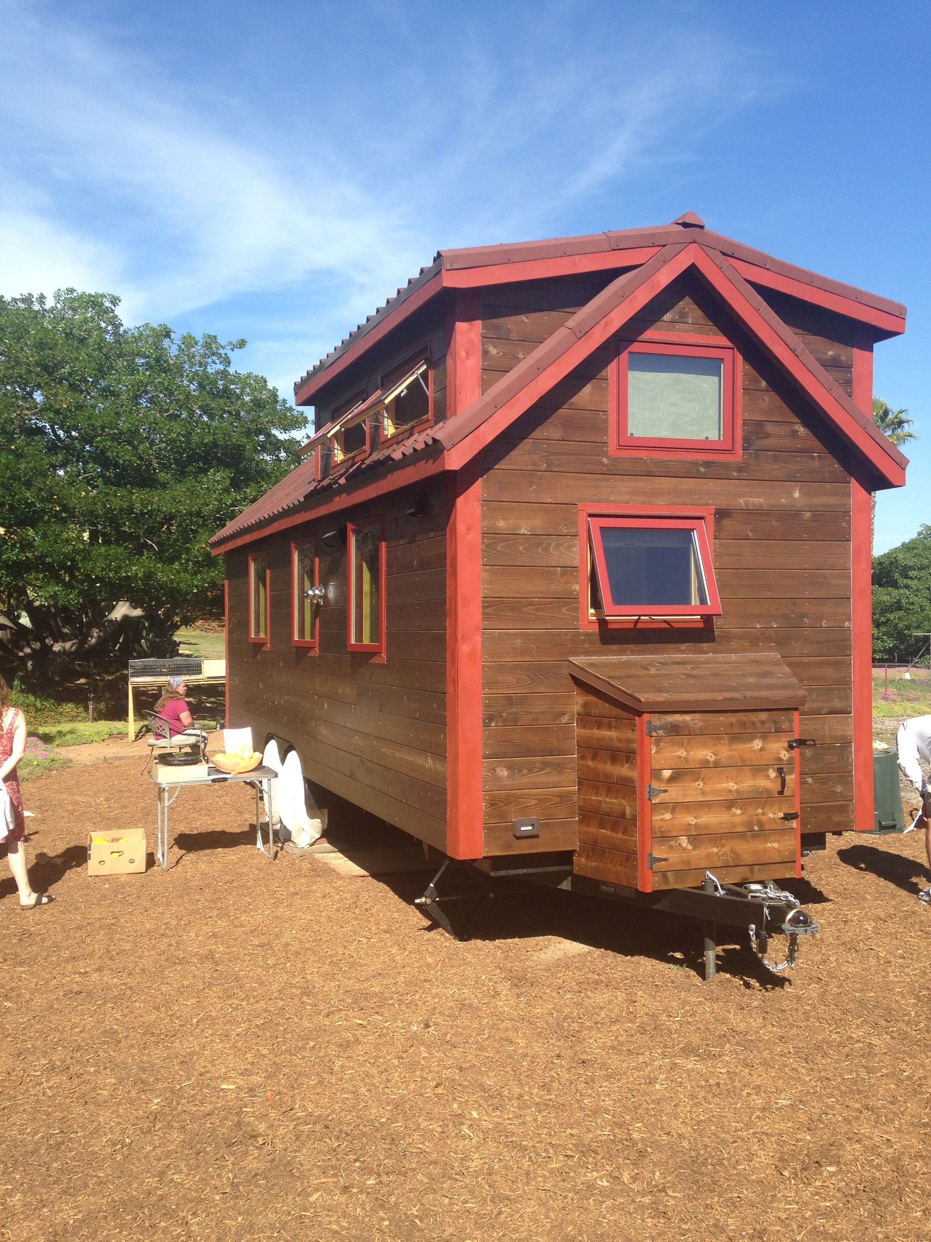 Tiny brown wooden house on wheels, with red trim, parked outdoors on wood chips under a blue sky.