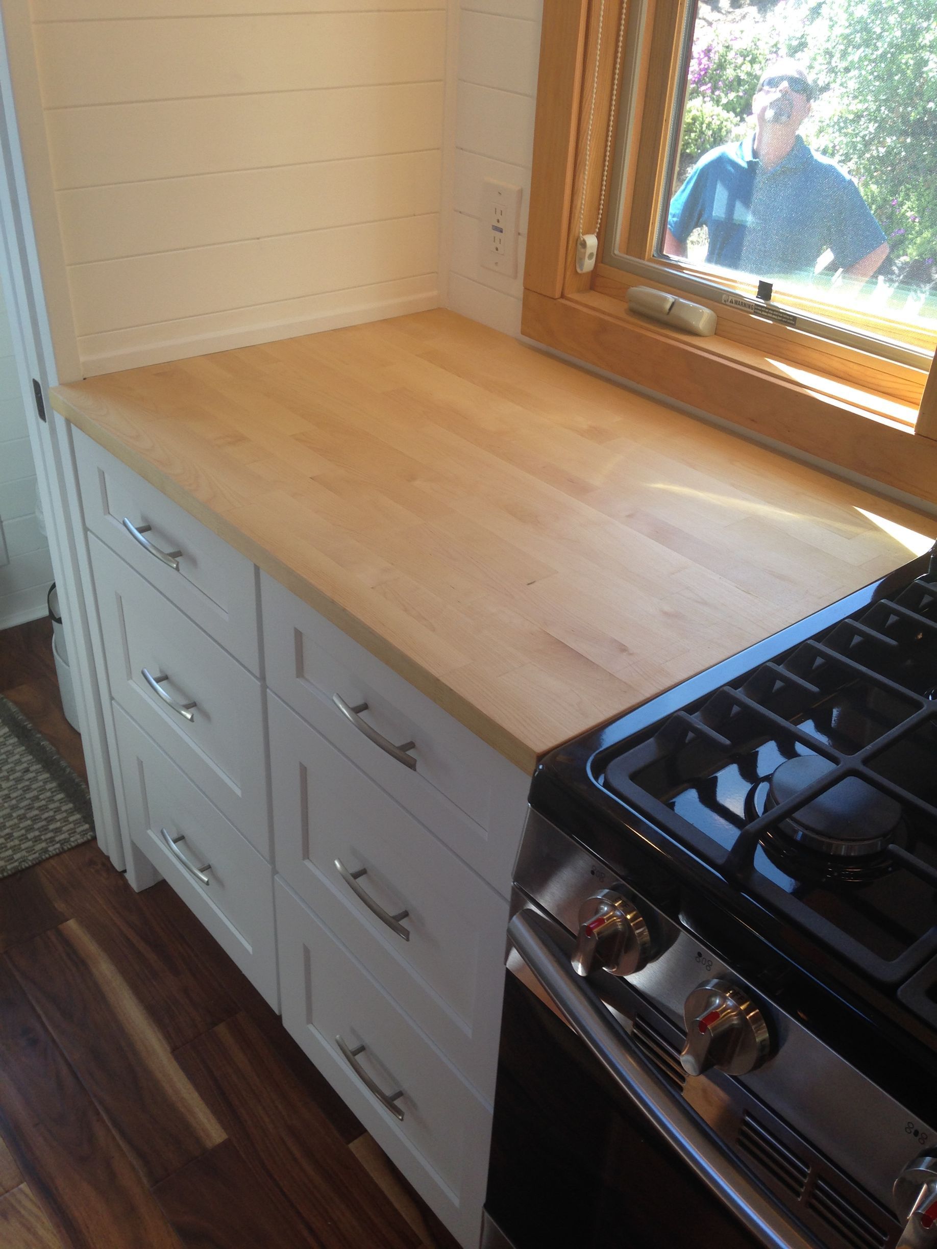 Kitchen with wooden countertop over white cabinets, stove on right, window with person in the background.