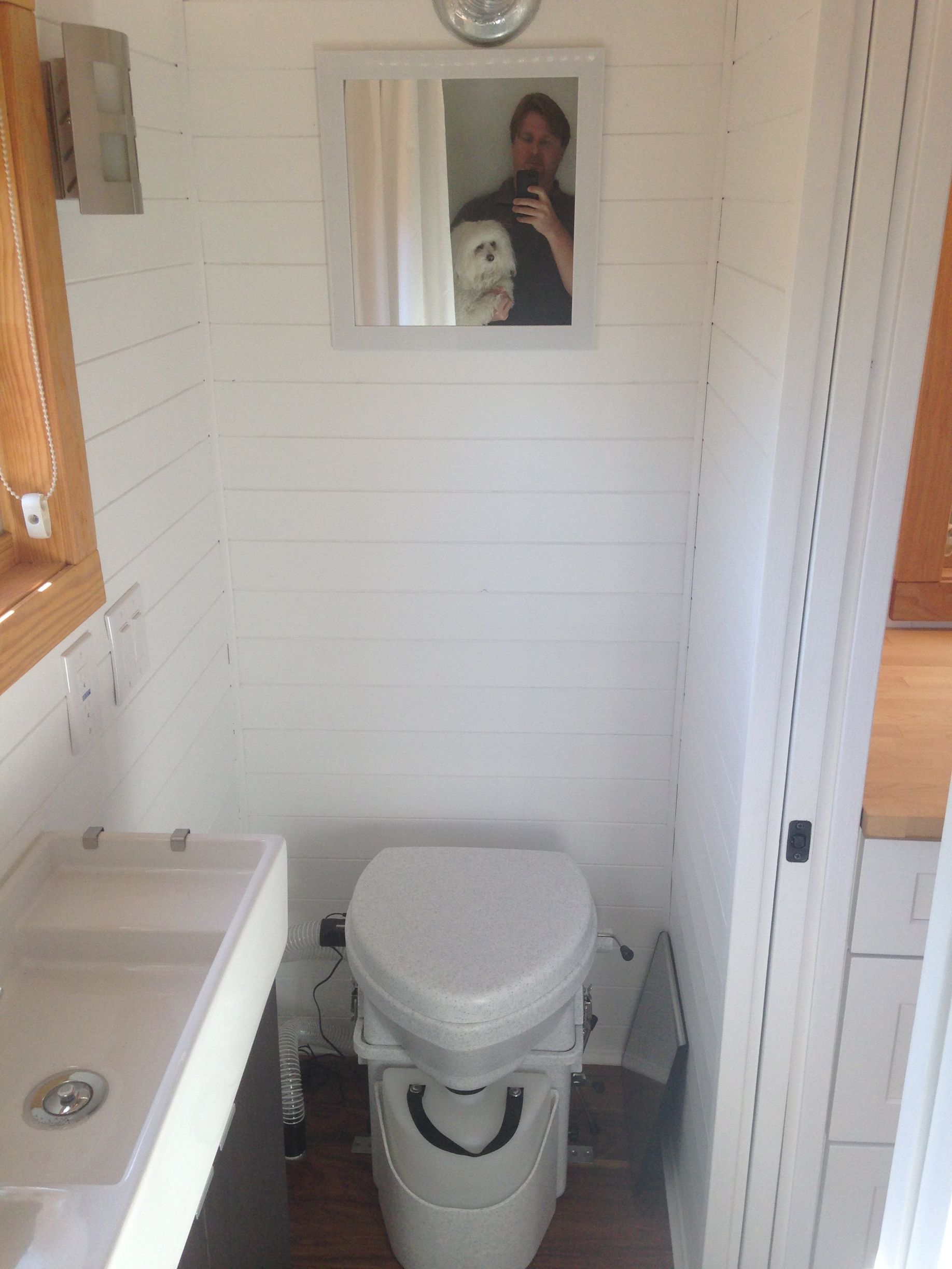 Small, white bathroom with a composting toilet, small sink, mirror reflecting person taking photo.