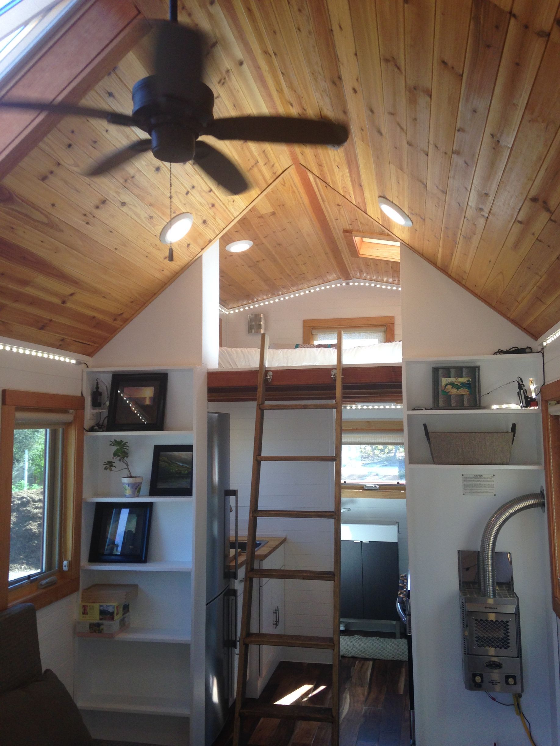 Interior of a tiny home with a loft, ladder, and built-in bookshelves. Wooden ceiling, natural light.