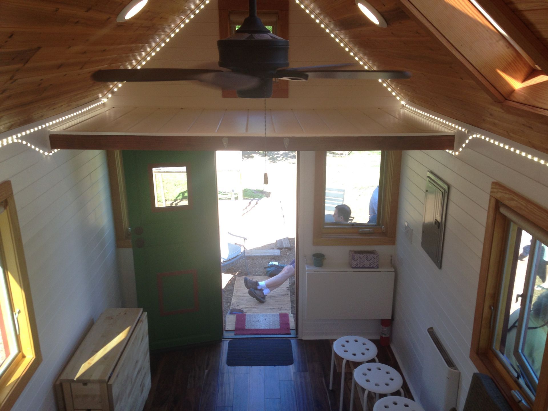 Interior view of a tiny home with open green door, ceiling fan, and small table with stools.