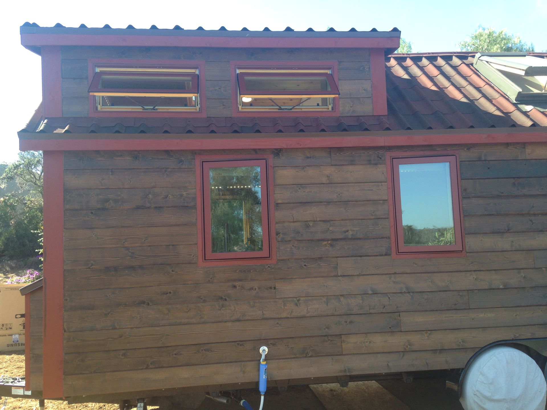 Tiny brown house with red trim and windows, on wheels, sunny outdoor setting.