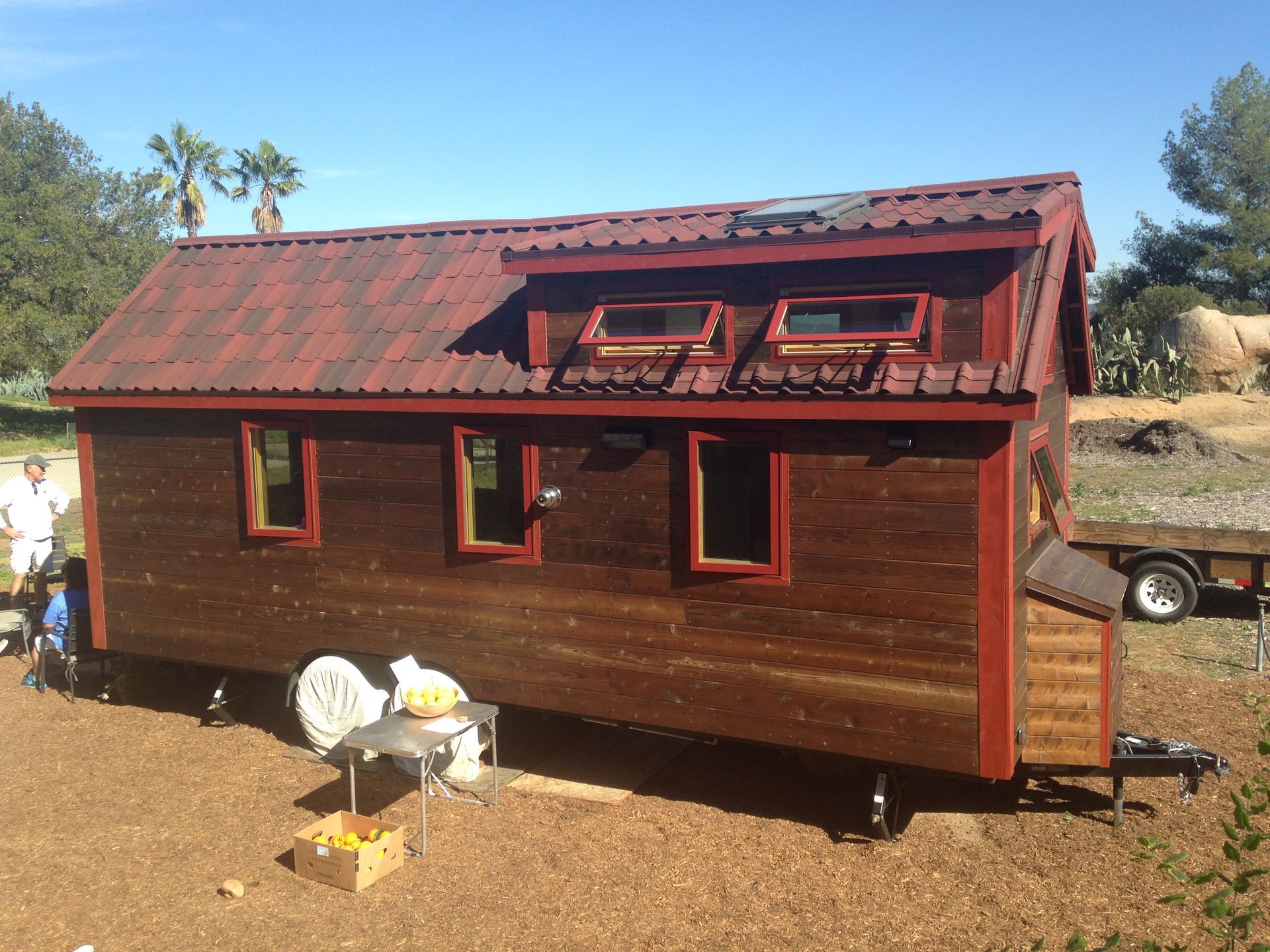 Tiny wooden house on wheels with red trim, on a sunny day.