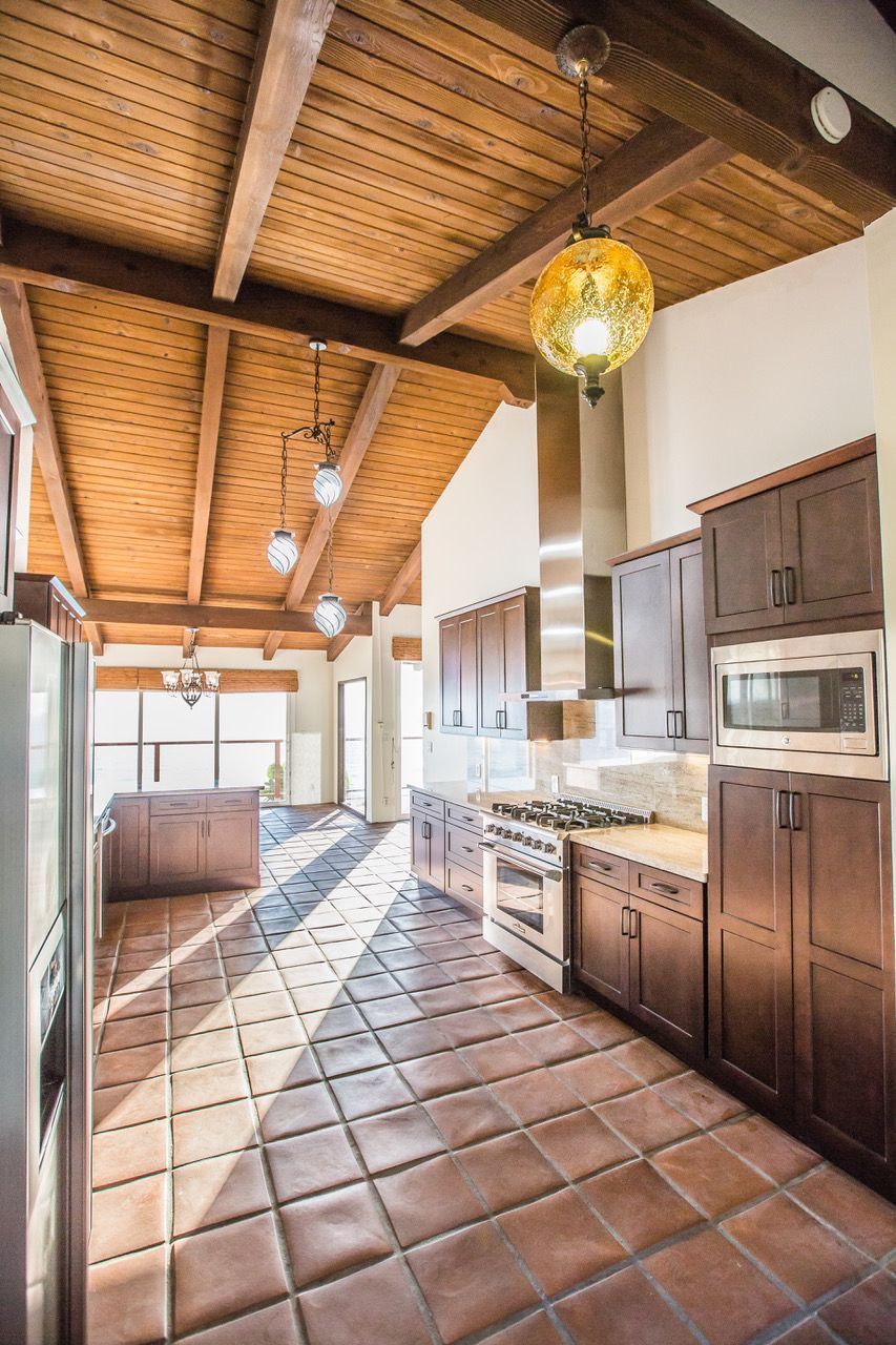 Kitchen with wood ceiling, brown cabinets, terracotta tile floor, and bright sunlight.