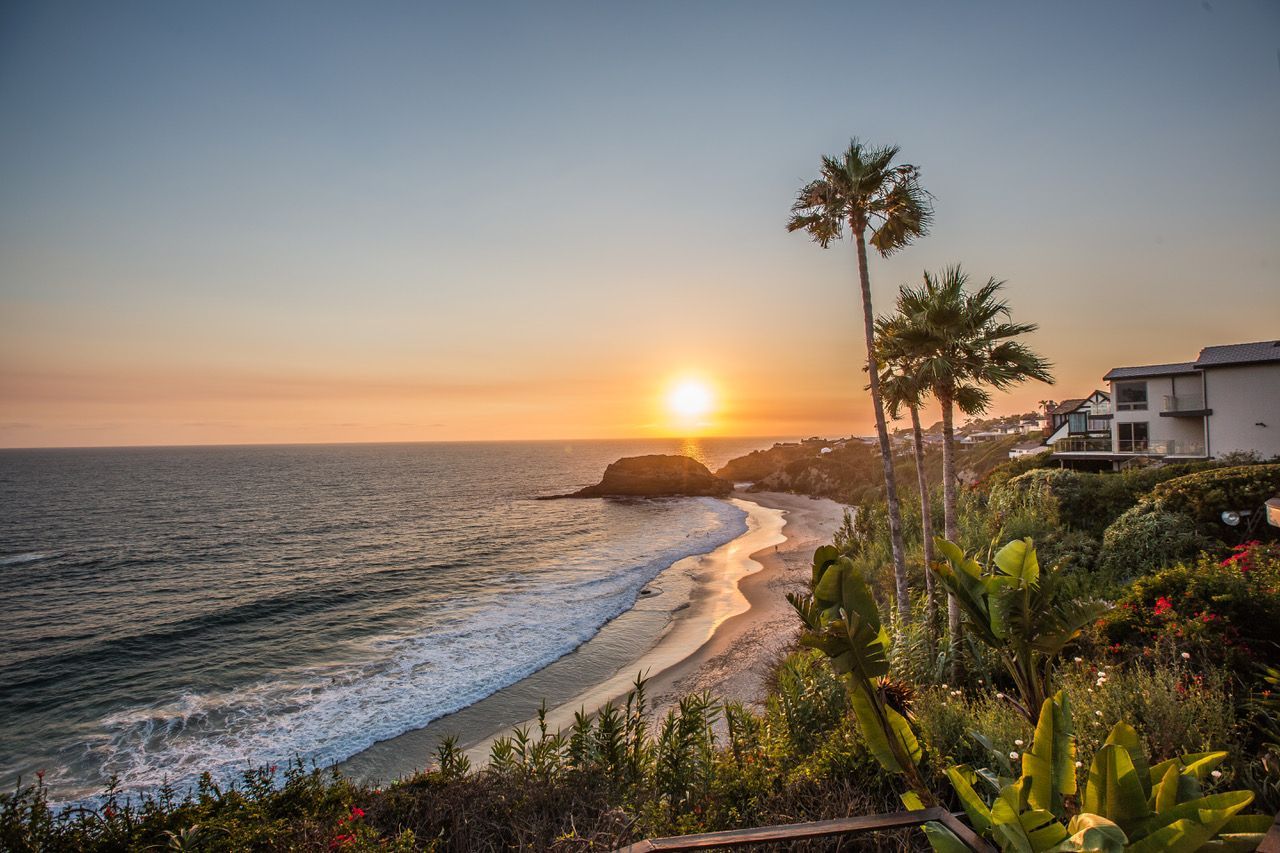 Sunset over a beach with palm trees, waves, and buildings in view; orange sky.