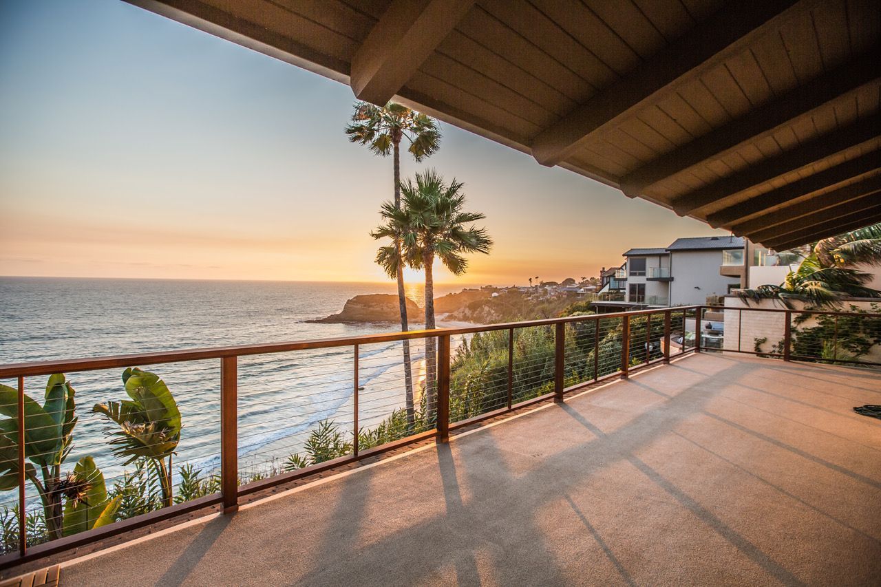 Ocean view from a balcony at sunset, with palm trees, wood railing, and building in the background.