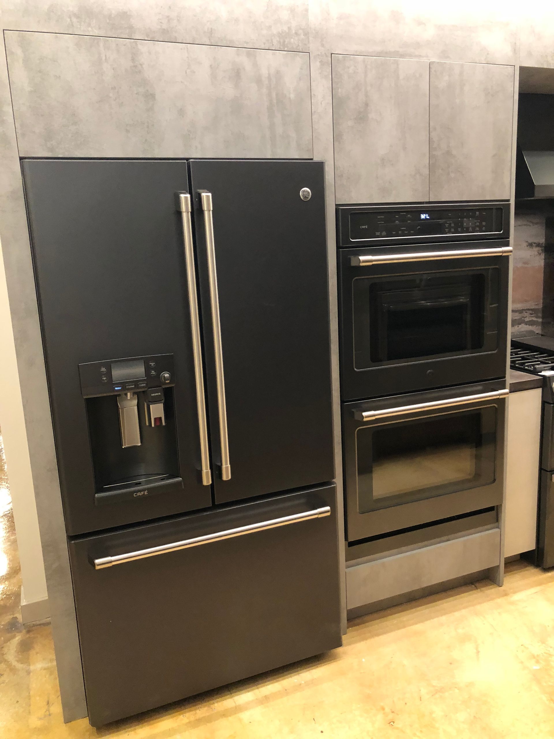 Black refrigerator and double oven in a gray kitchen setting.
