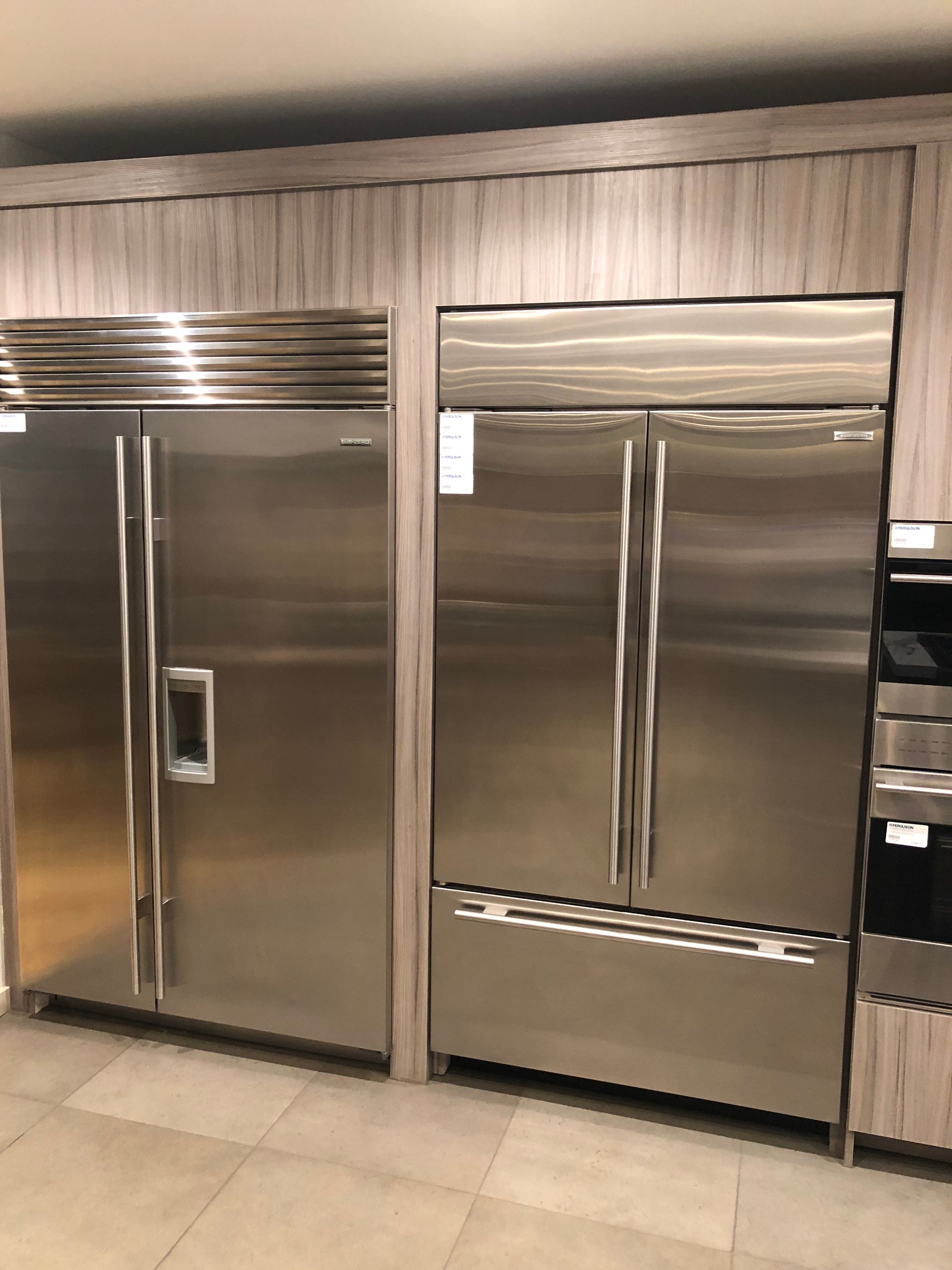 Stainless steel refrigerators side-by-side, built into a wooden cabinet in a showroom.