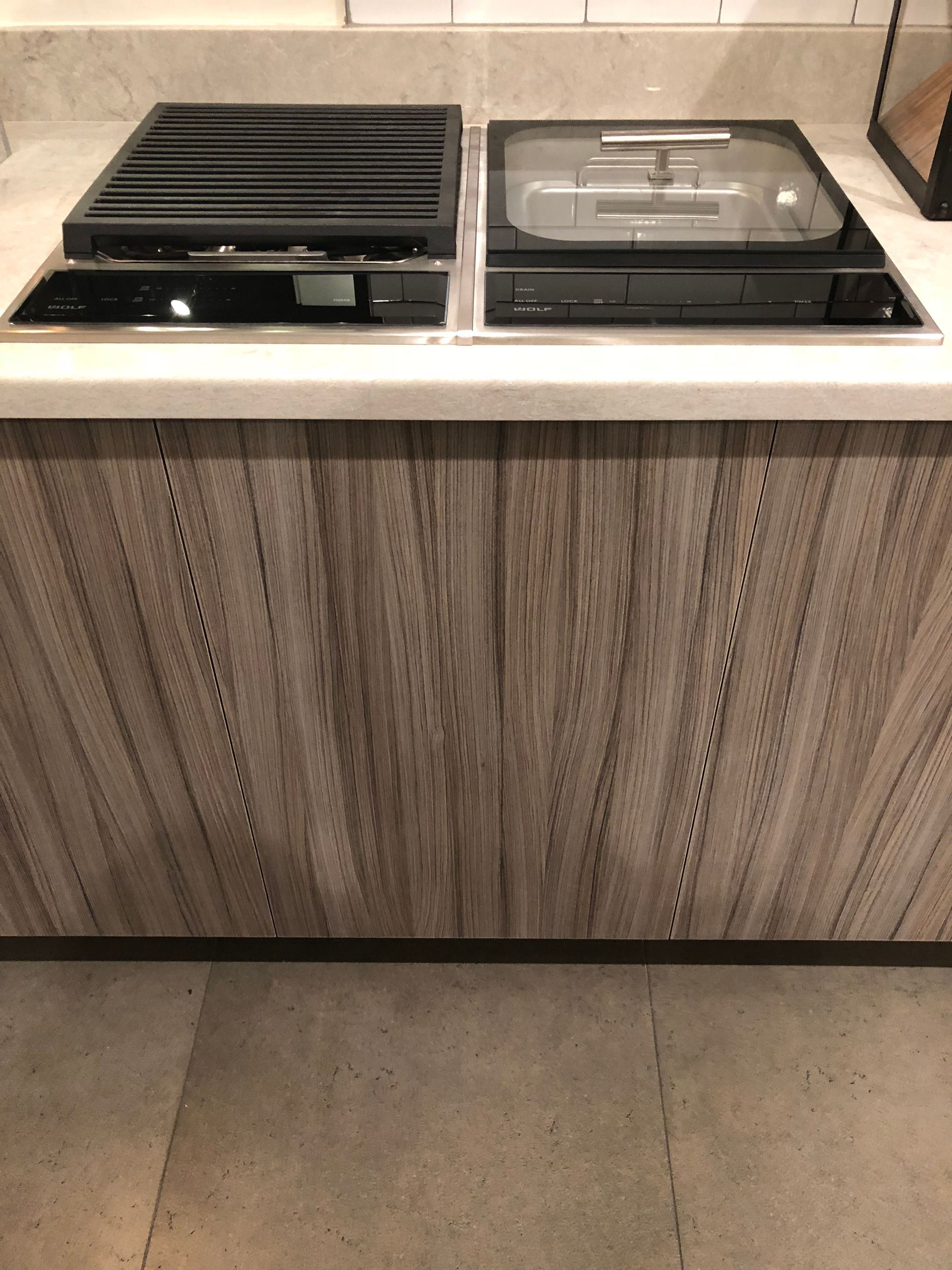Kitchen countertop with a grill and cooktop, over light wood grain cabinet doors and a tiled floor.