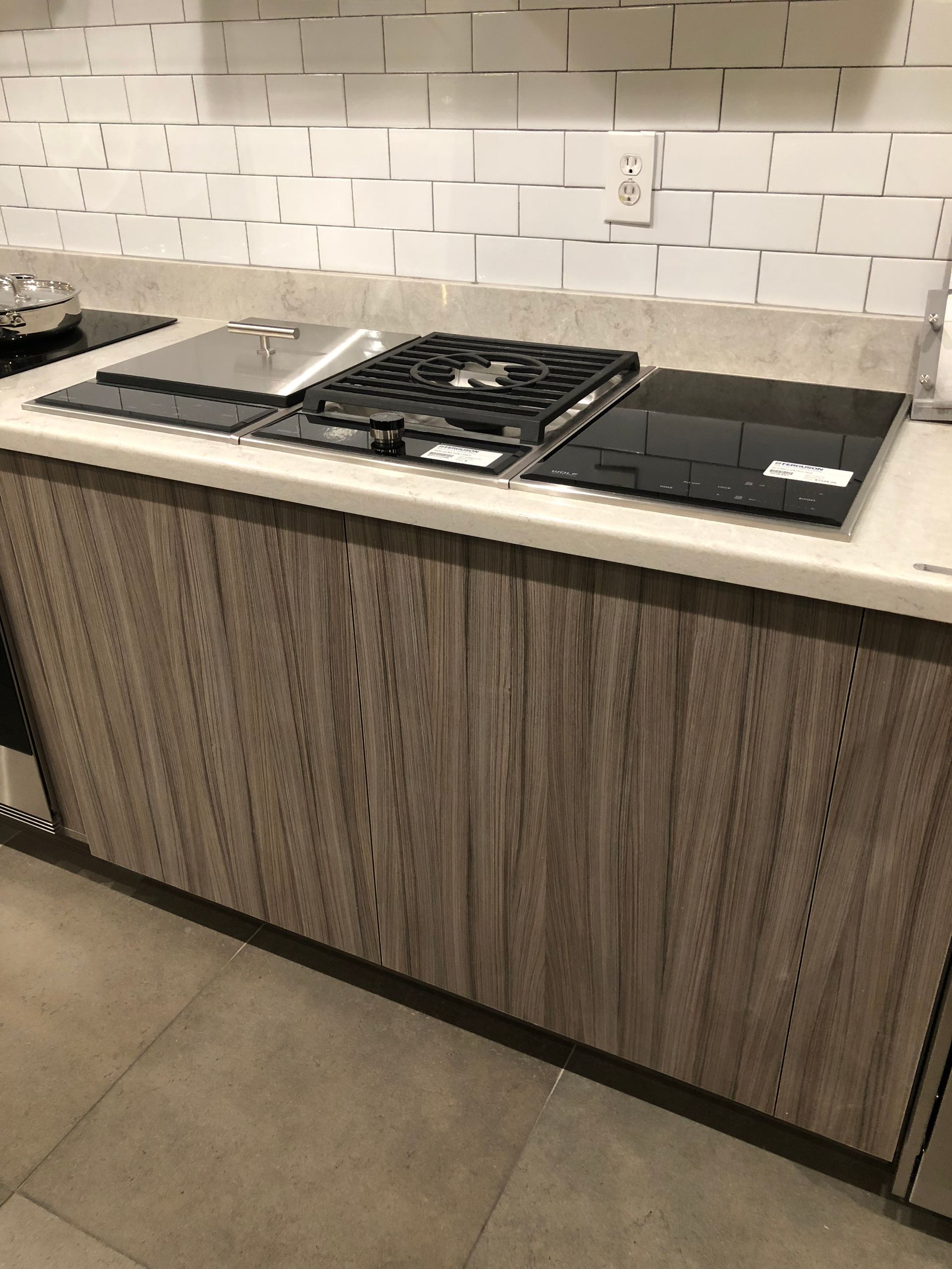 Kitchen counter with stove top and wood-grain cabinets. White subway tile backsplash, neutral-toned countertops.
