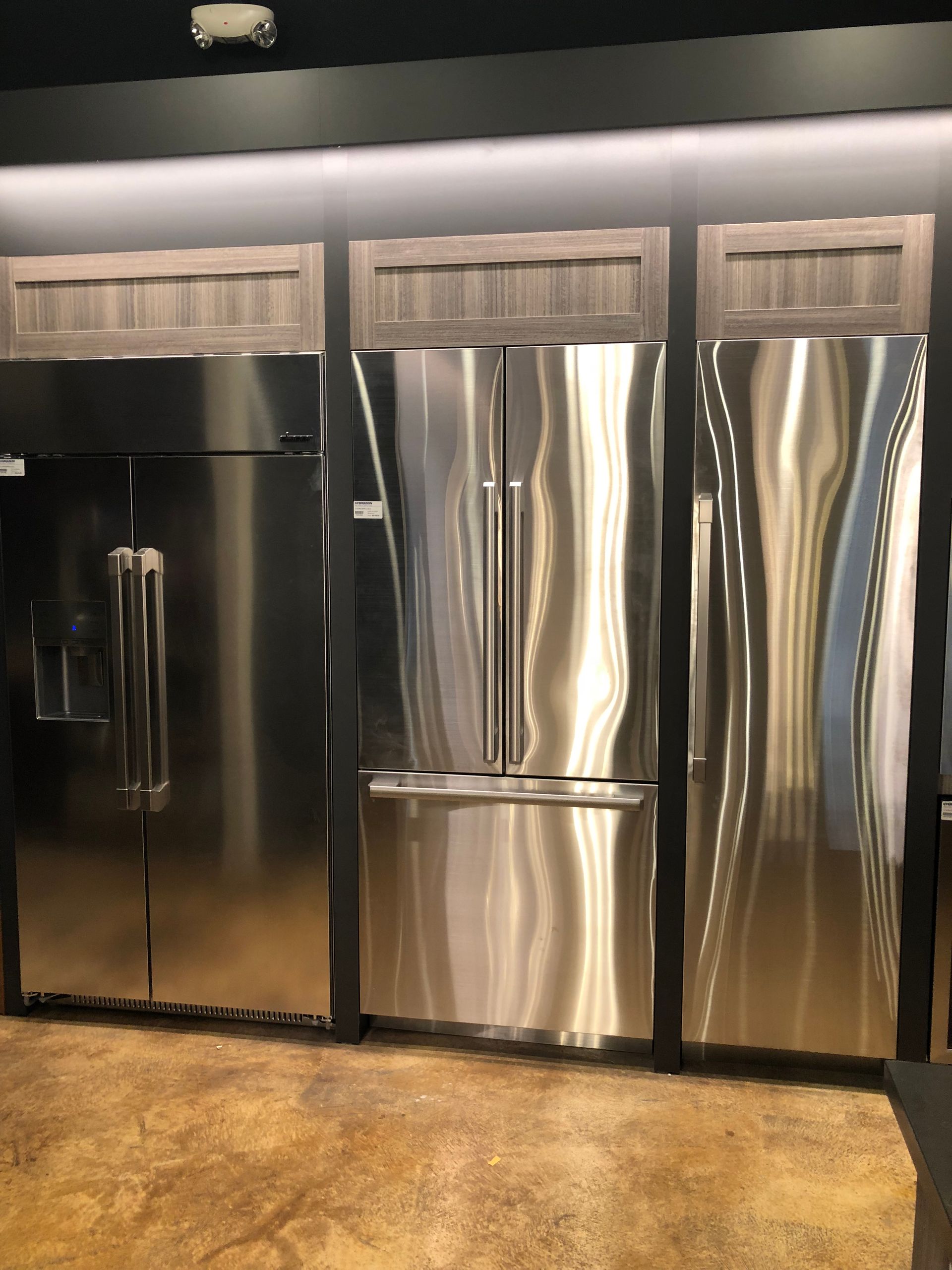 Three stainless steel refrigerators displayed, wooden cabinets above, on a brown floor.