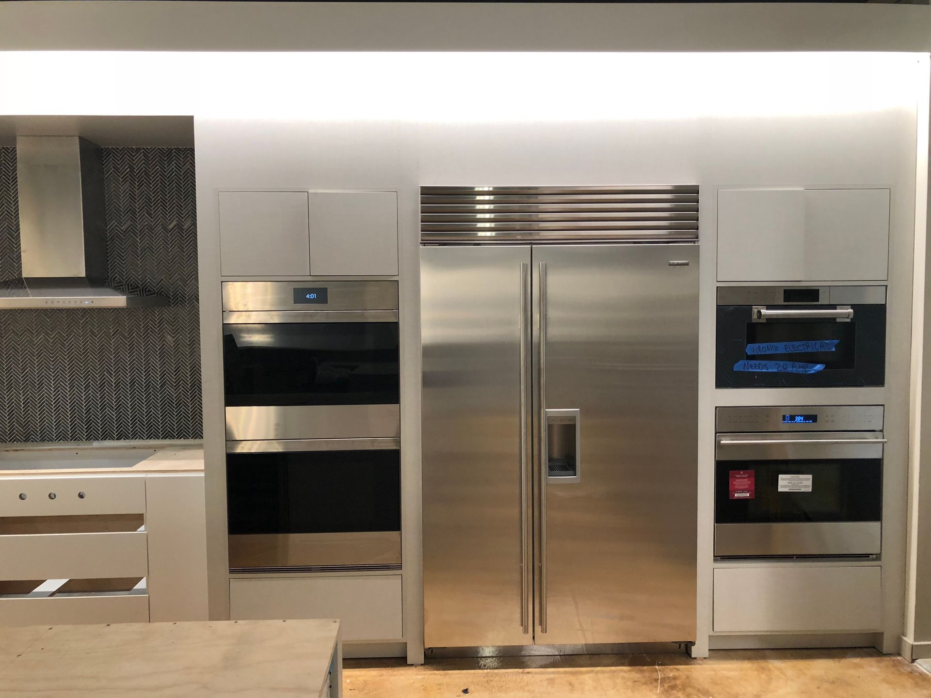 Stainless steel appliances in a modern kitchen: refrigerator, ovens, and range hood. White cabinets and recessed lighting.
