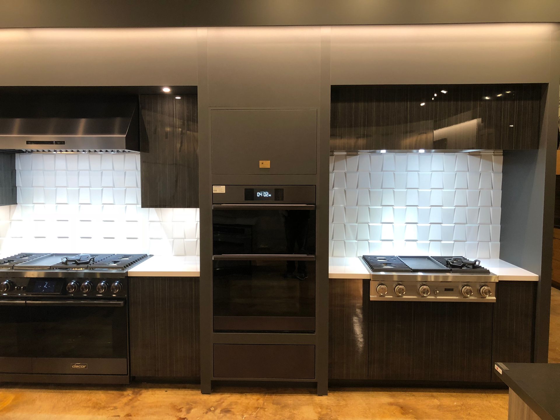 Kitchen display with dark cabinets, ovens, stoves, and white tile backsplashes.