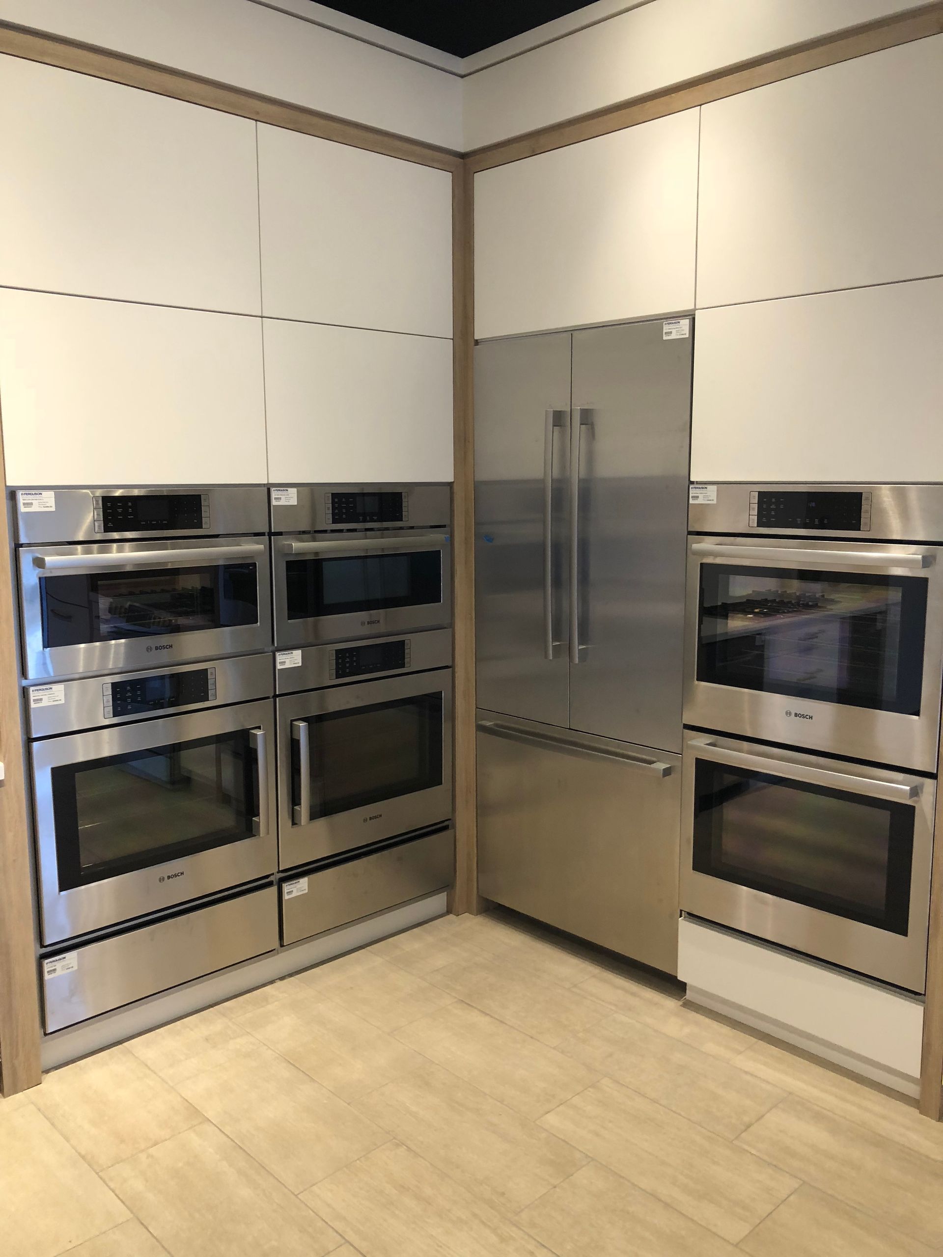 Kitchen appliances display: stainless steel ovens, refrigerator, and white cabinets. Beige flooring and wood trim.