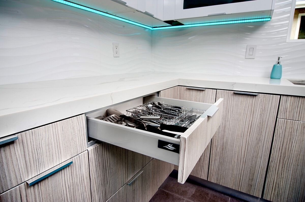 A kitchen with an open drawer filled with silverware. Beige cabinets, white countertop, and blue accent lighting.