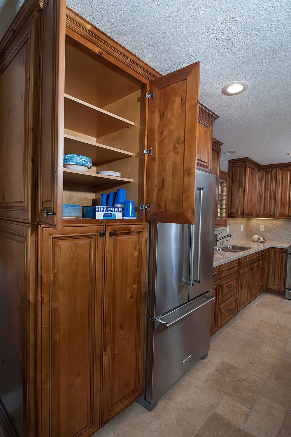 Tall wooden kitchen cabinet with open door, stainless steel refrigerator, and shelves holding blue items.