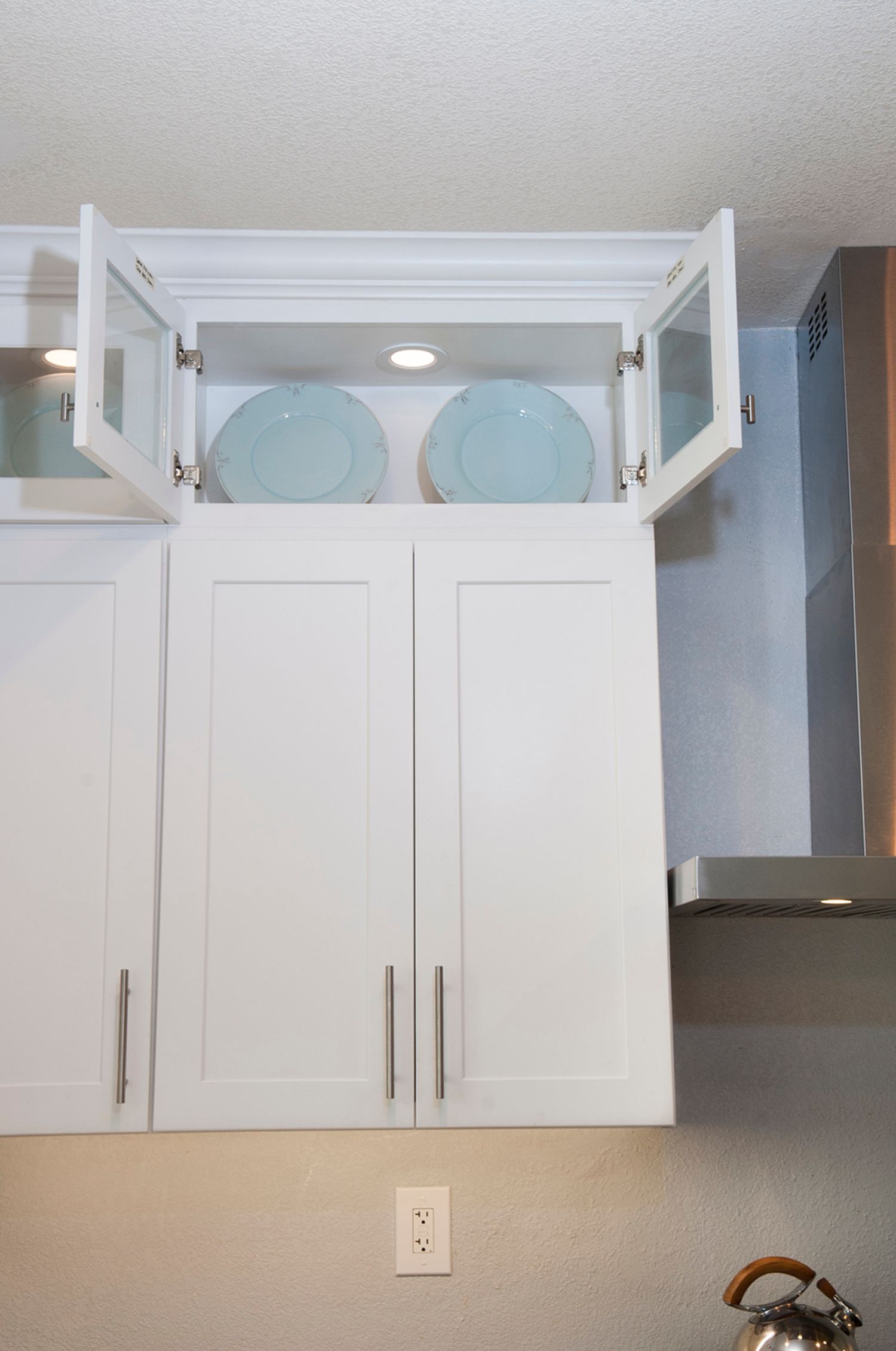 White kitchen cabinets with glass-door top cabinet holding blue plates, range hood on the right.