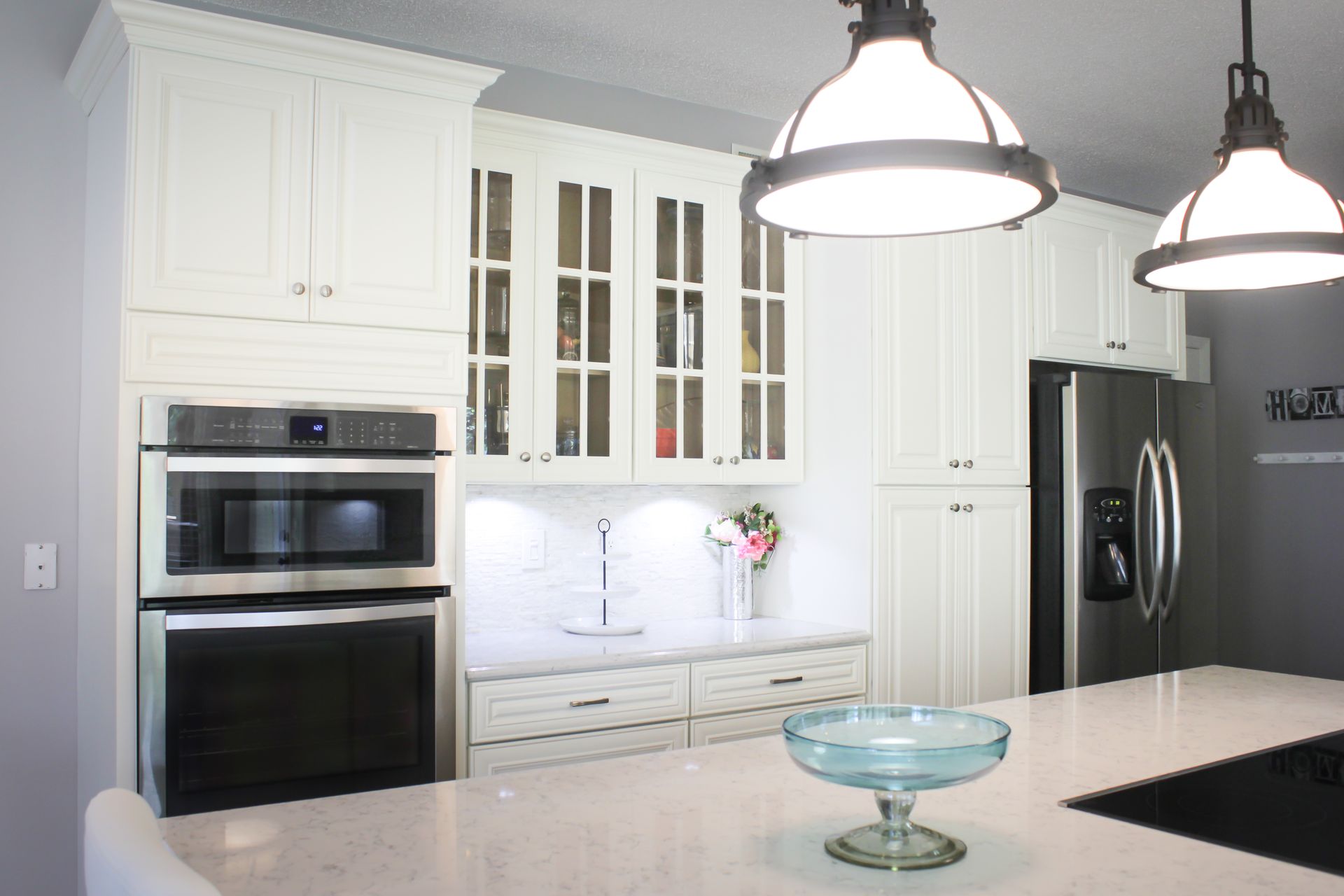 White kitchen cabinets with a built-in oven and microwave, stainless steel refrigerator, and pendant lights.