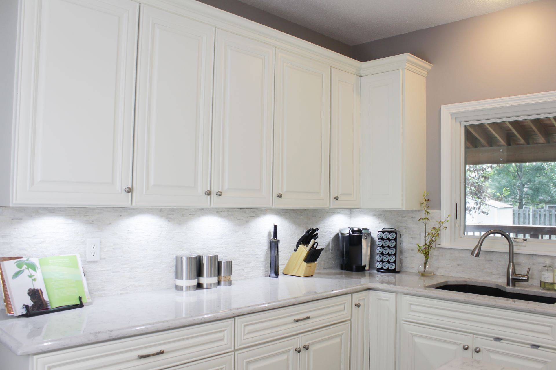 White kitchen cabinets, countertops, and backsplash with under-cabinet lighting and a window.