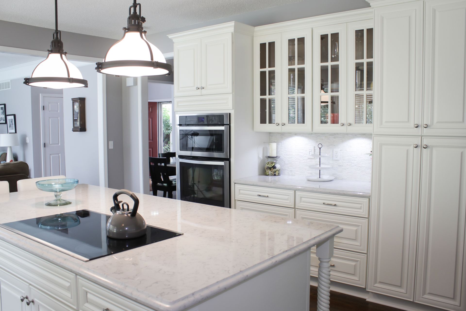 White kitchen with island, cabinets, and built-in oven. Black pendant lights hang above.