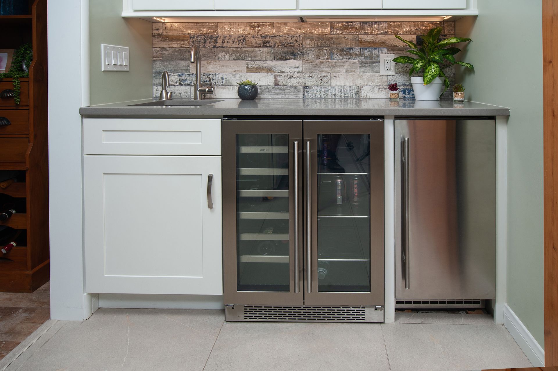 Small home bar with white cabinets, stainless steel fridge, and wine cooler. Gray countertop and decorative backsplash.