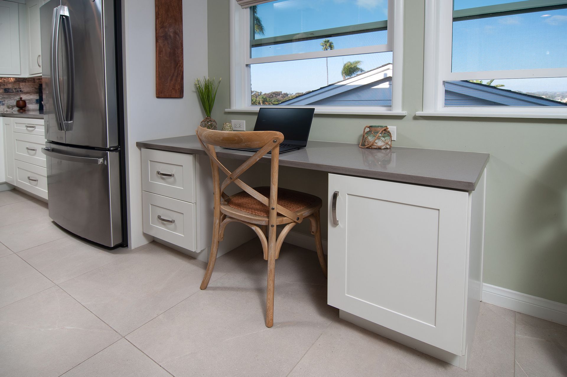 A home office nook in a kitchen with a desk, chair, and laptop by a window, with a refrigerator to the left.