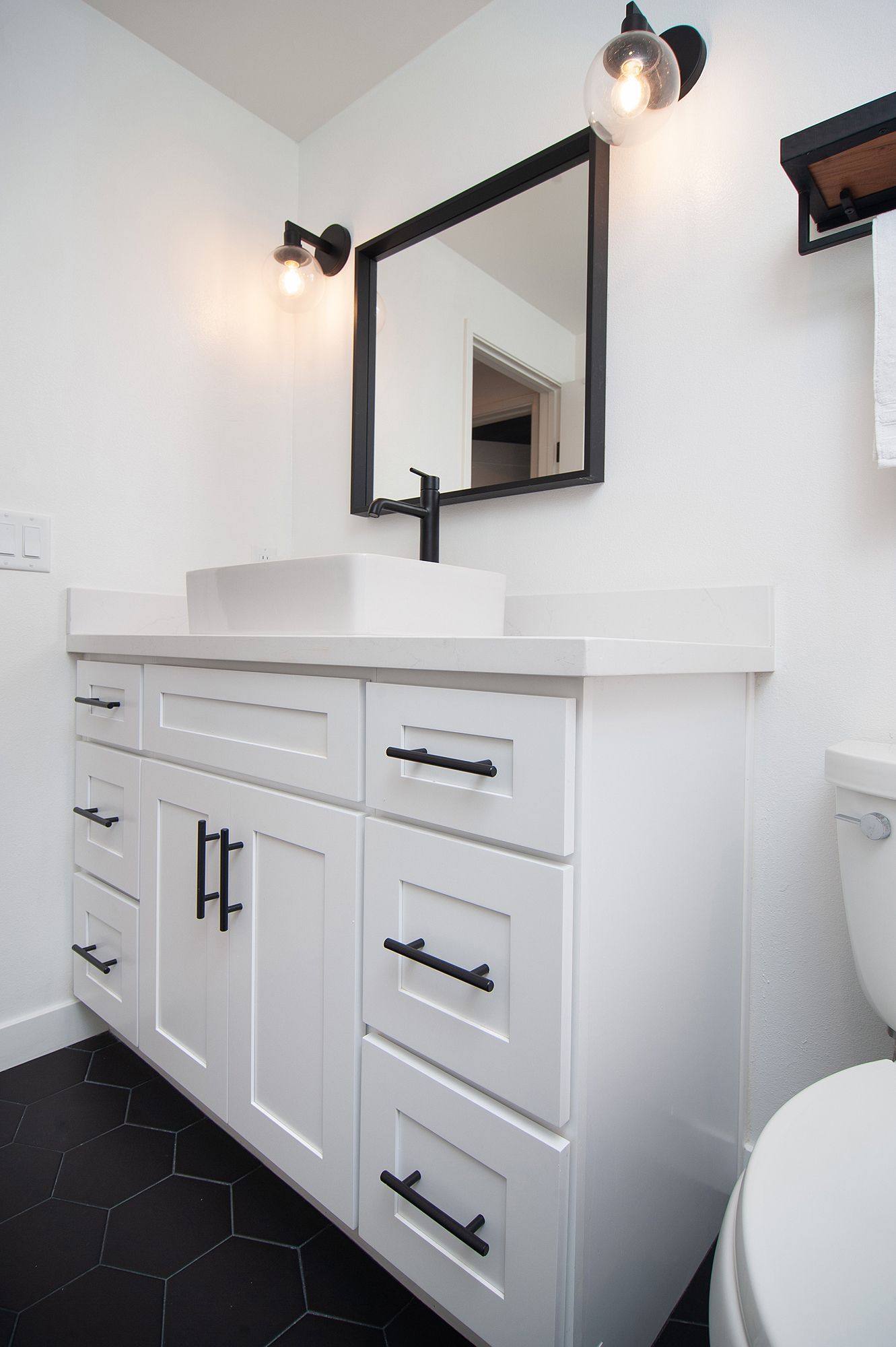 White bathroom vanity with black accents, mirror, and lights. Black hexagonal floor tiles.