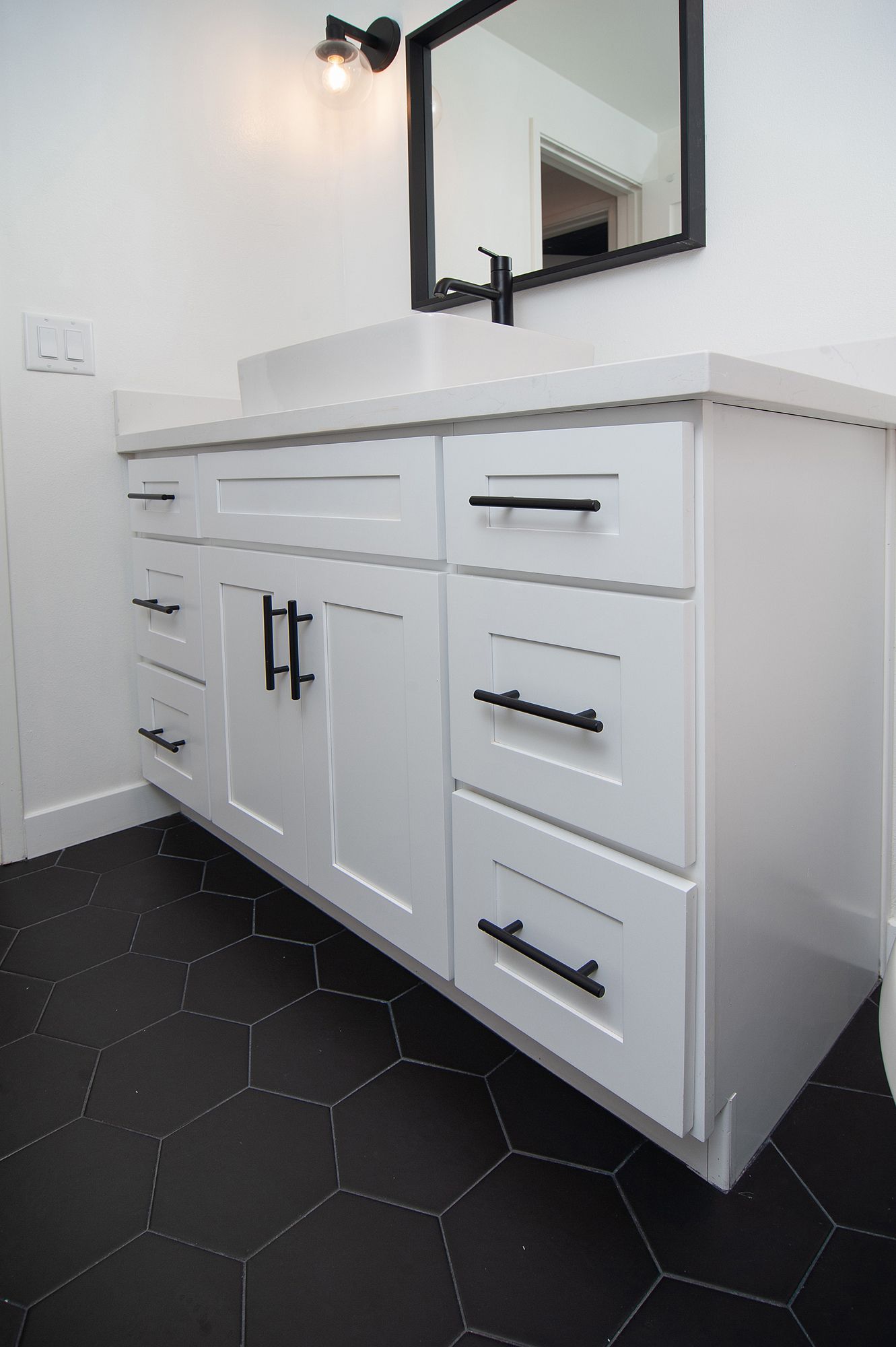 White bathroom vanity with black hardware, black hexagon tile floor, and a mirror.