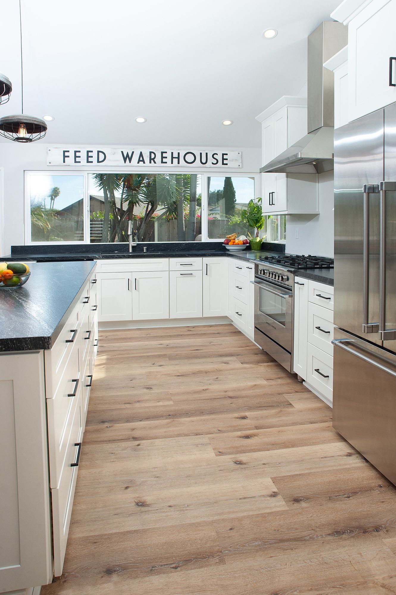 Modern white kitchen with dark countertops, stainless steel appliances, and wood-look flooring.