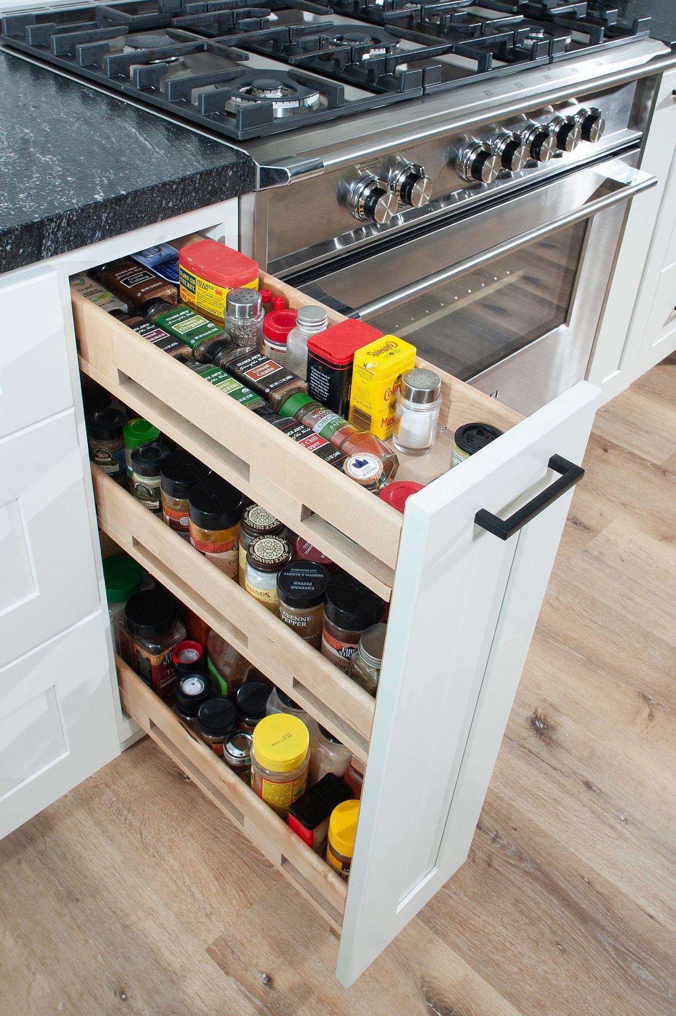 Open spice rack beside a stainless steel oven, filled with various spice jars and containers. White cabinet, wood floor.