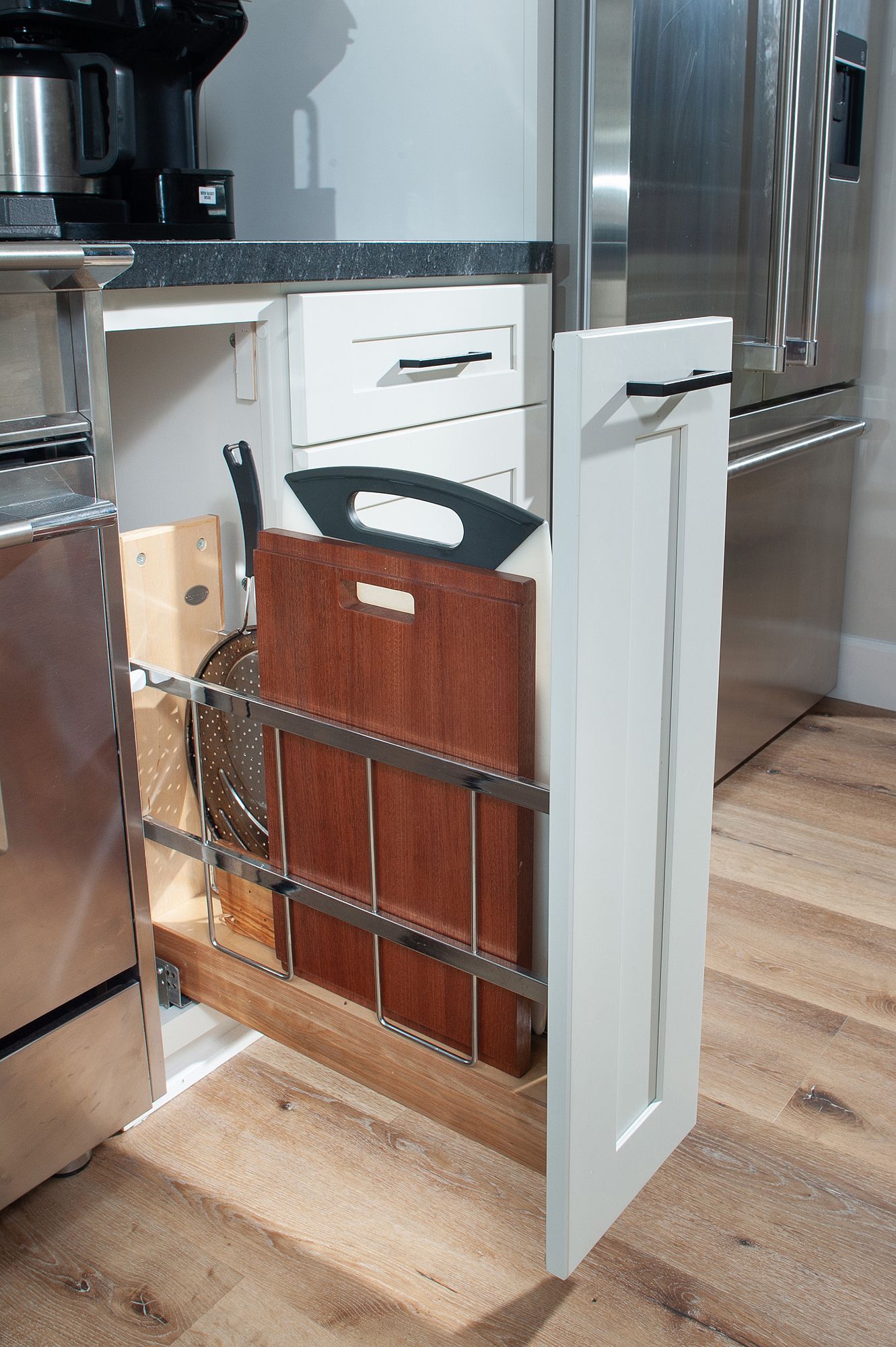 White kitchen cabinet with pull-out storage for cutting boards and pans, next to a stainless steel appliance.
