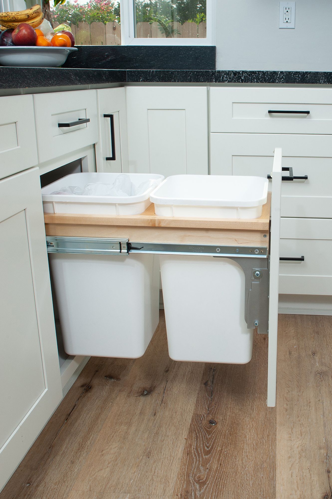 White kitchen cabinet with pull-out trash bins; two white receptacles on a wooden pull-out tray.